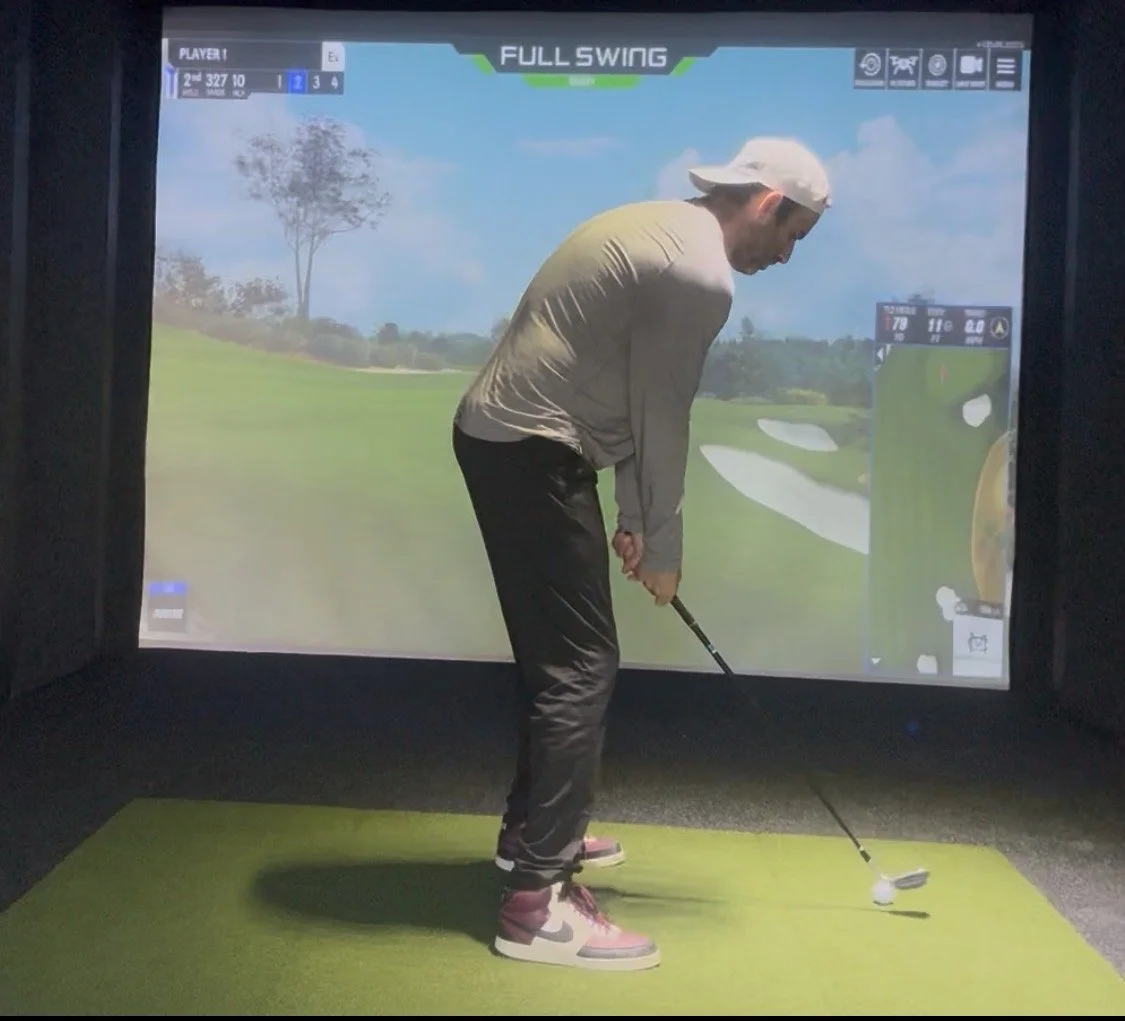 A person practicing golf indoors on a screen with a golf simulator, preparing to hit a golf ball on a green course.