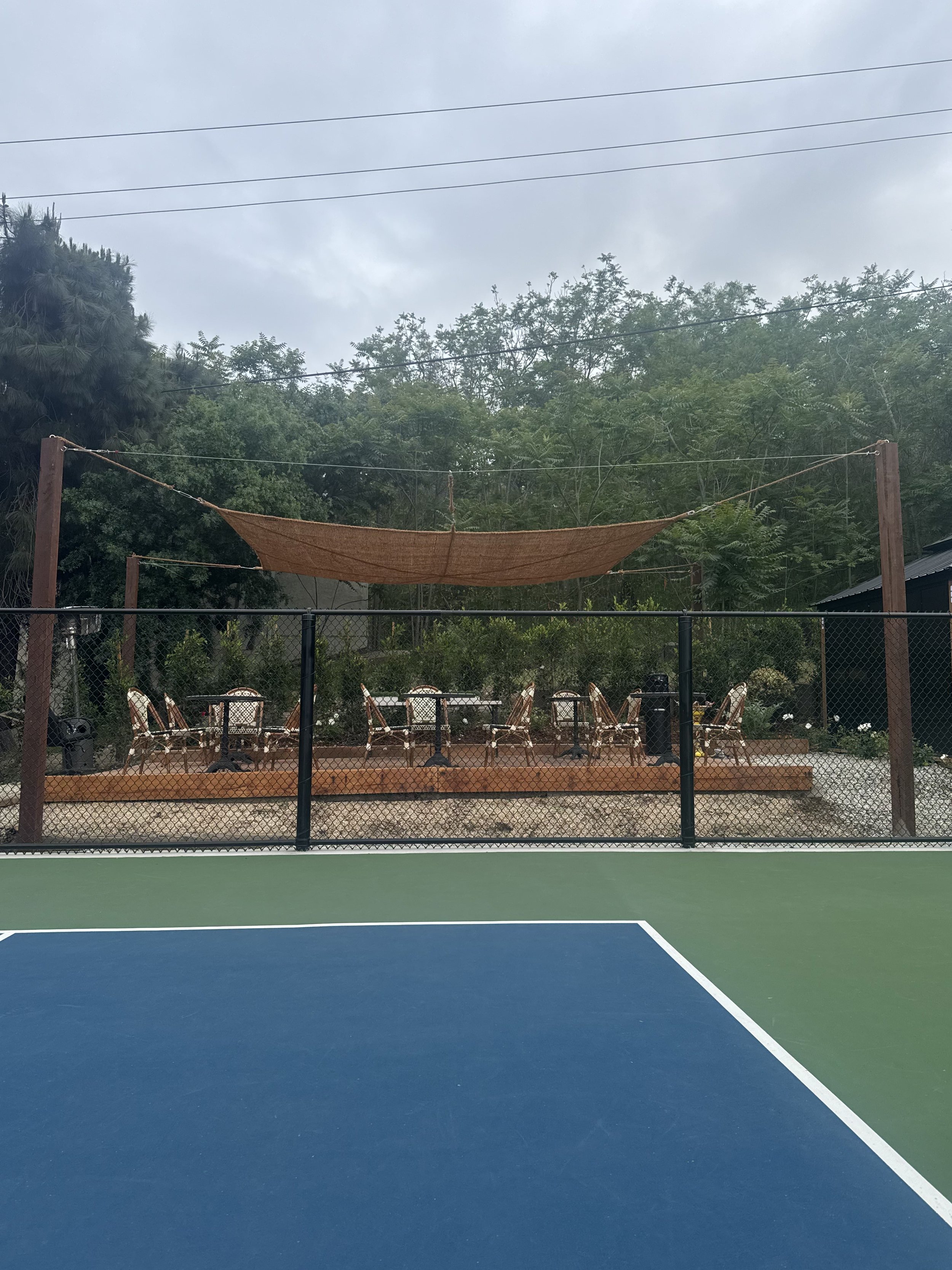 View of an outdoor restaurant with seating area featuring white chairs with brown frames, shaded by a brown fabric canopy, situated behind a sports court with blue and green surface and black fencing. Trees and overcast sky in the background.