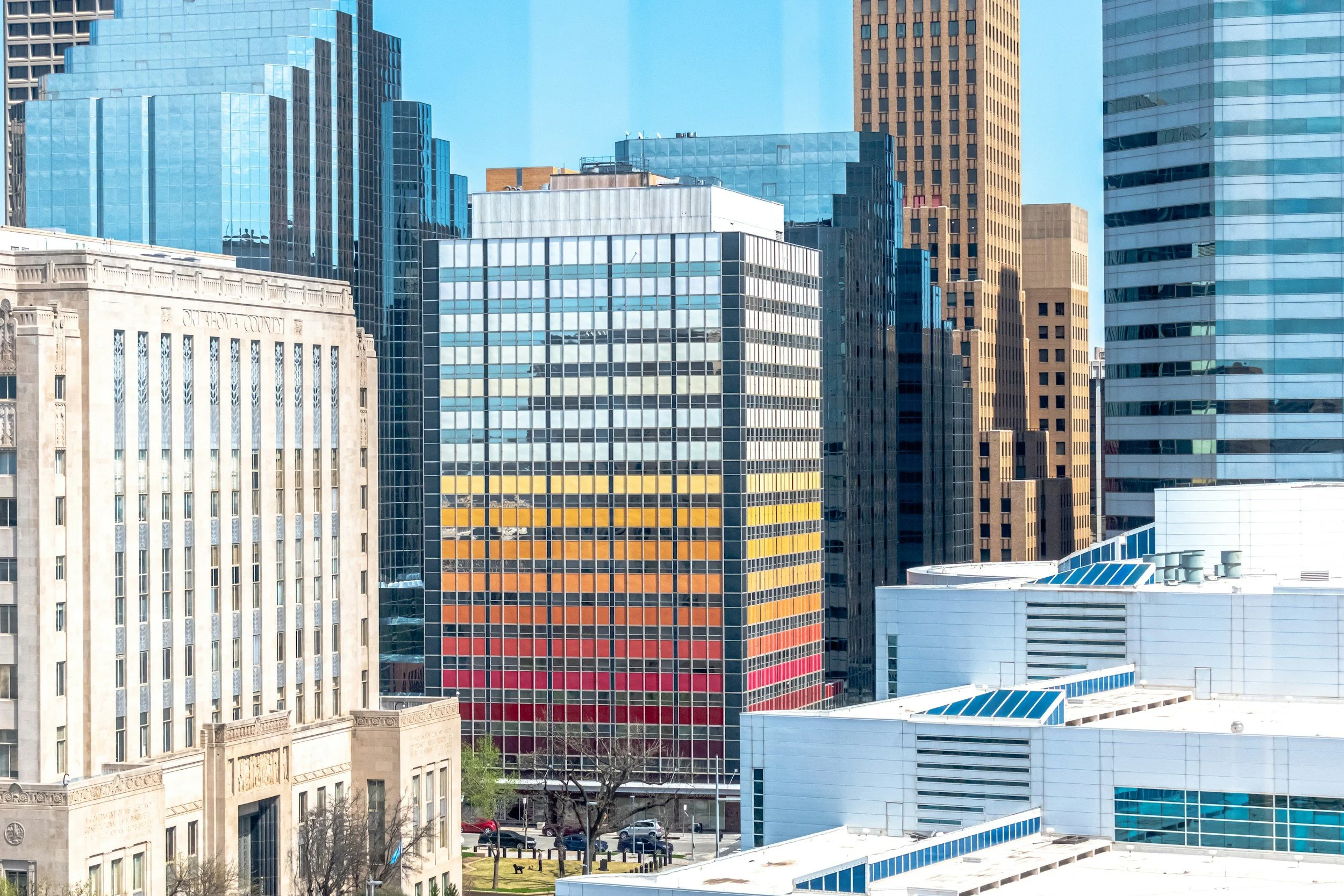 Cityscape with tall modern buildings, including a glass office tower with colored stripes, and a historic stone building in the foreground.
