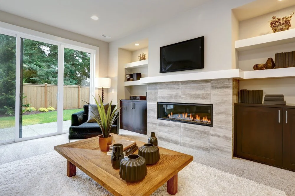 Modern living room with fireplace, wall-mounted TV, sliding glass door, armchair, and wooden coffee table with decorative vases.