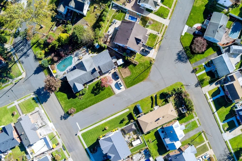 An aerial view of a residential neighborhood with houses, driveways, lawns, trees, and backyard swimming pool.