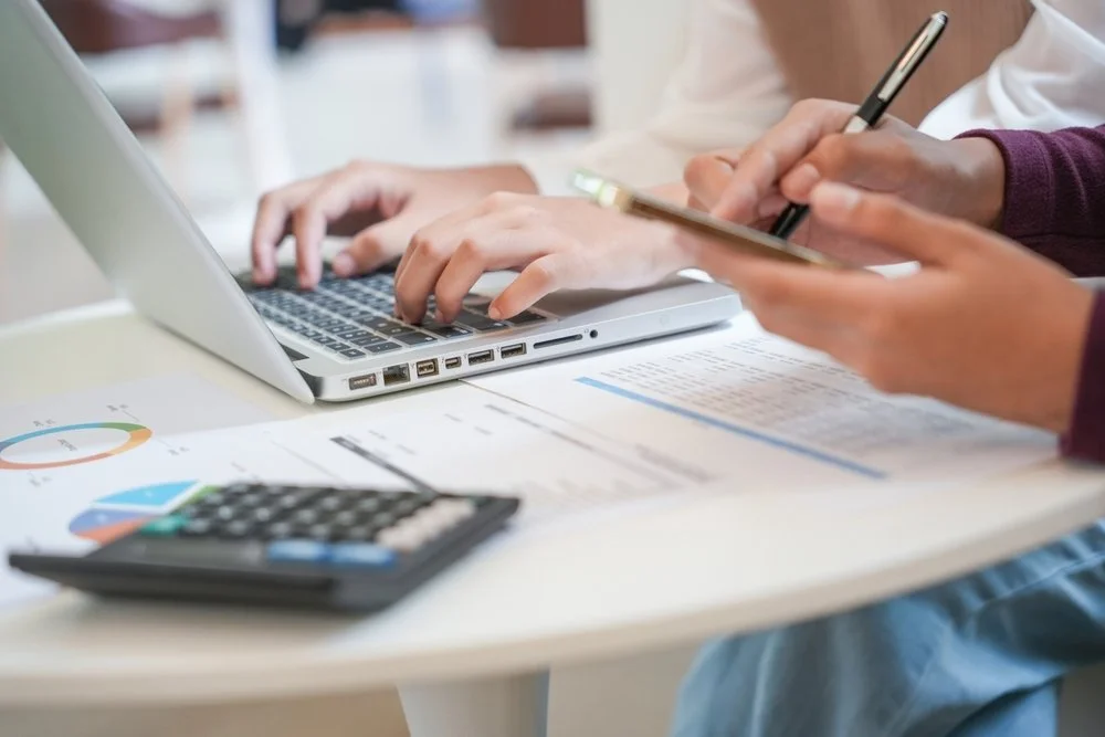 People working at a desk with a laptop, calculator, documents, and a person using a smartphone and taking notes.