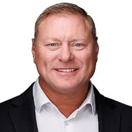 A smiling man wearing a black suit jacket and white shirt against a plain background.