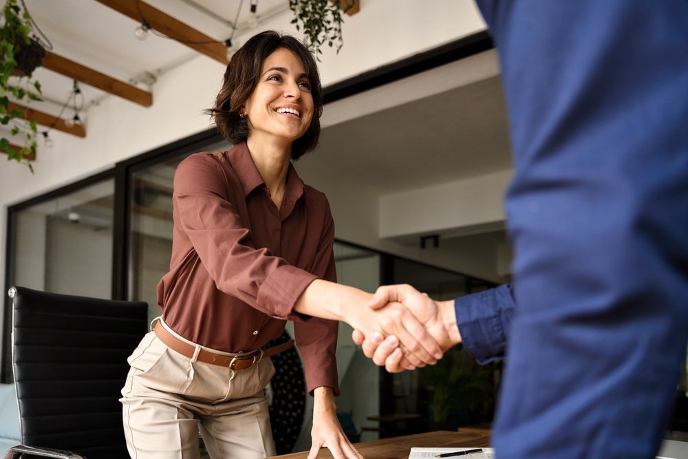 Woman smiling and shaking hands with a man in a modern office setting.