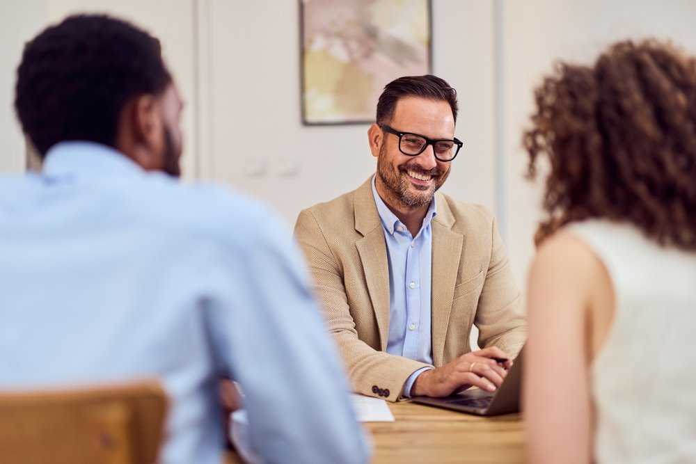 A man in glasses and a tan blazer is smiling during a meeting with two other people at a table.