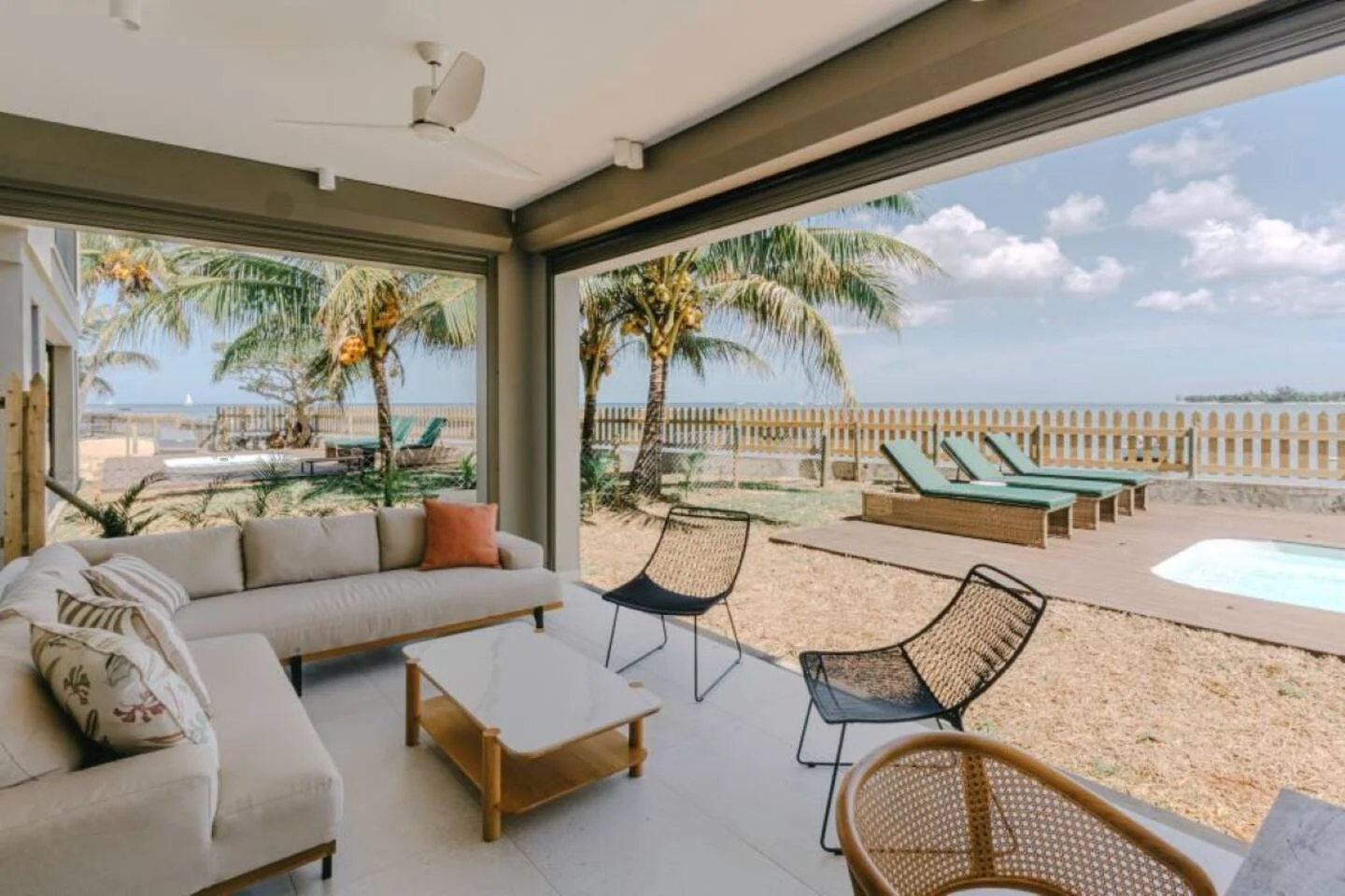 View from the interior of a beach house living room looking out to a sandy yard with lounge chairs, palm trees, a fence, a small pool, and the ocean in the background under a partly cloudy sky.