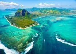 Aerial view of a tropical island surrounded by turquoise waters and coral reefs, with lush green hills and a sandy beach. Showing where Mauritius whale swim is