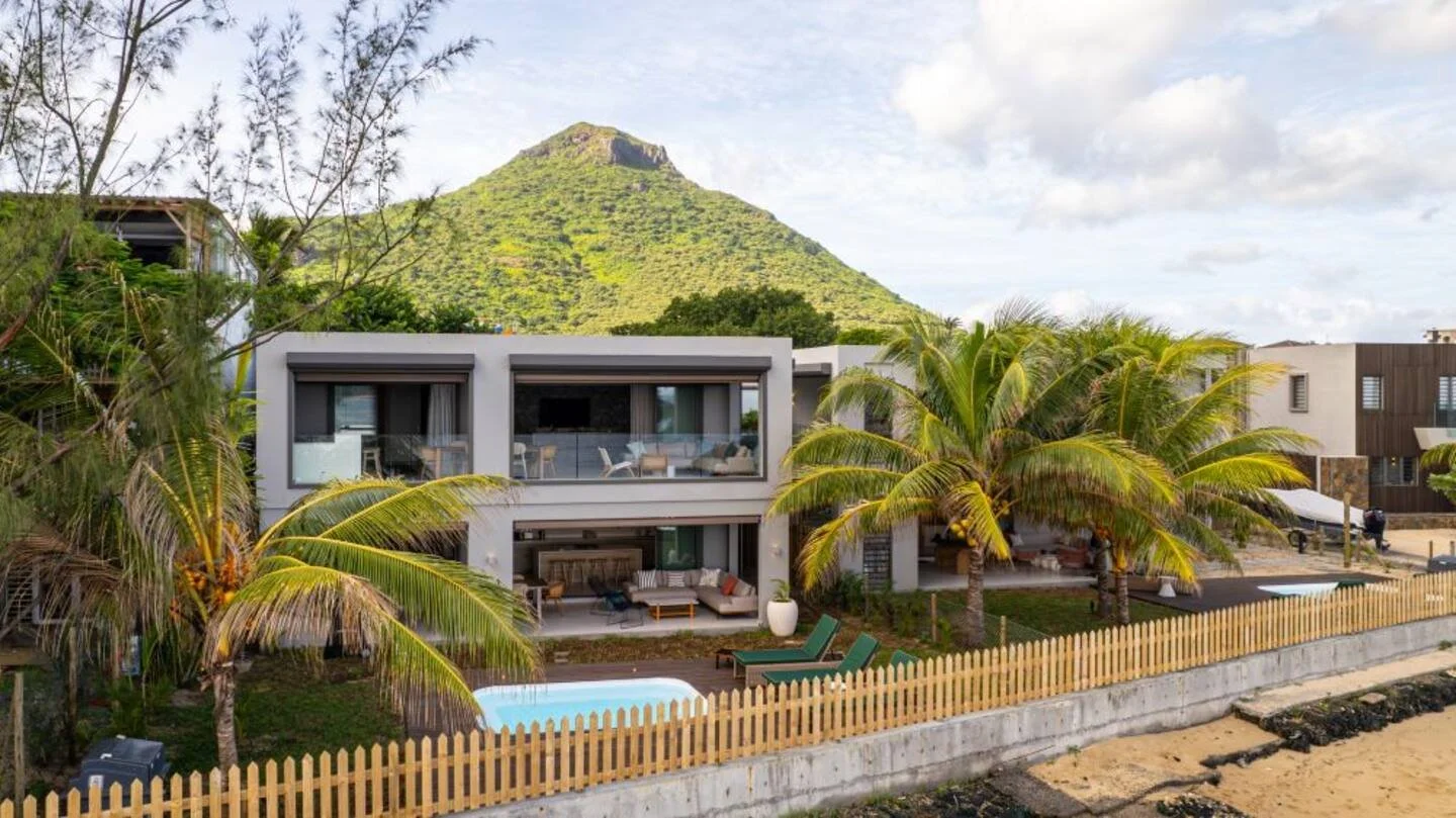 Modern two-story Mauritius house with large windows, pool, and lounge chairs in a tropical setting with palm trees and a mountain in the background.
