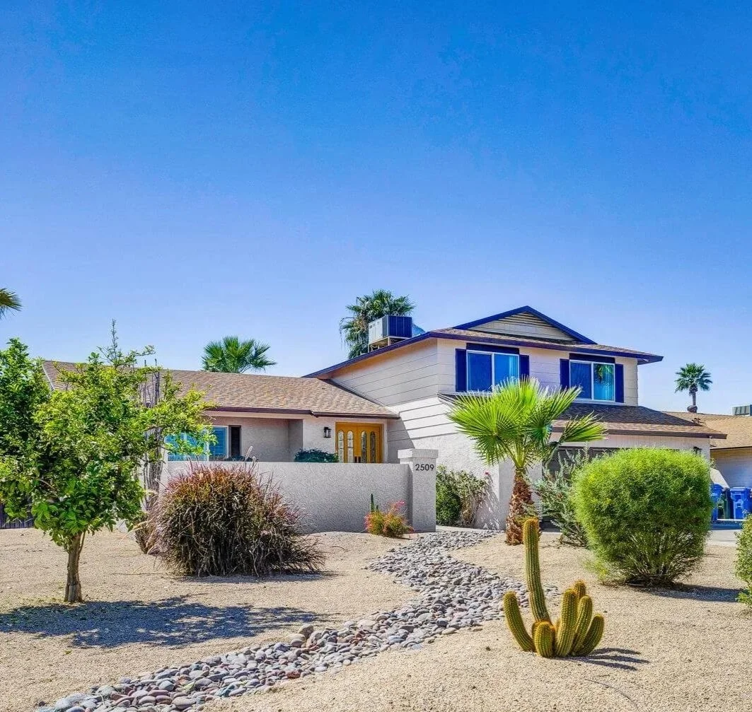 A bright wide-angle shot of a beige two-story home. The yard is covered in tan gravel, surrounded by drought-tolerant plants and small cacti under a clear, cloudless blue sky.