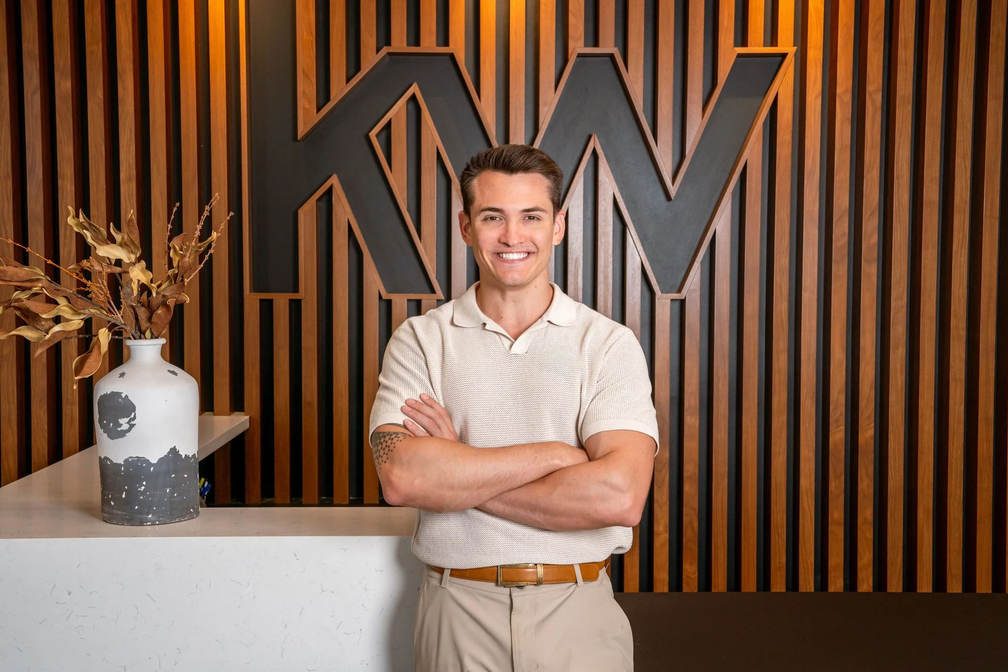 Ryan Zamudio, a young man with short brown hair and a light beard, smiling with arms crossed, standing in front of a wooden wall with a large geometric letter M behind him.