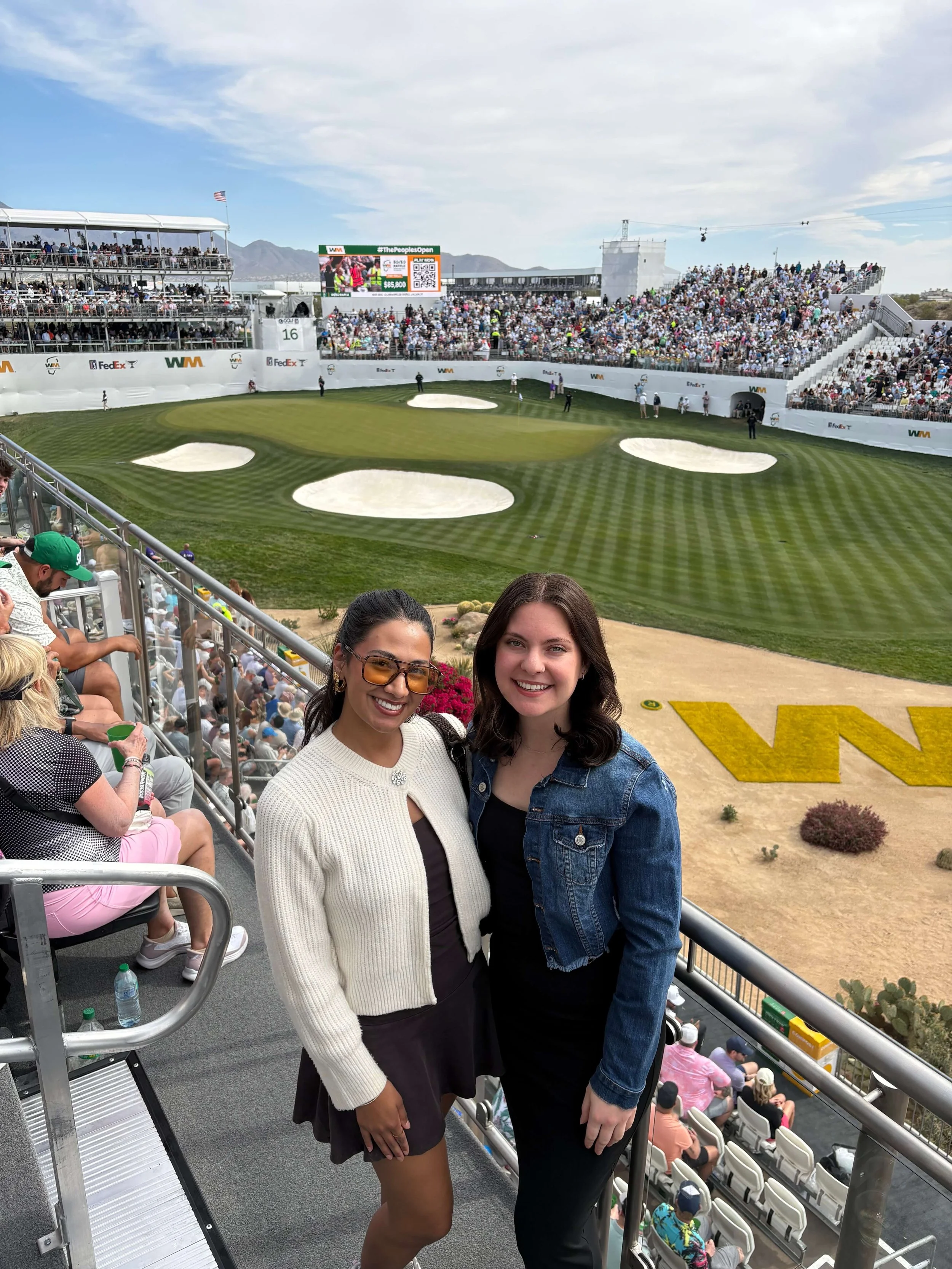 Two members of the Bryant Real Estate team smiling at the famous 16th hole during the WM Phoenix Open golf tournament in Scottsdale, Arizona.