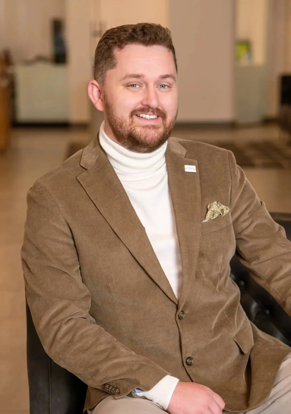 Professional headshot of Scott Bryant, luxury real estate agent, wearing a brown corduroy blazer and white turtleneck in a modern office setting.