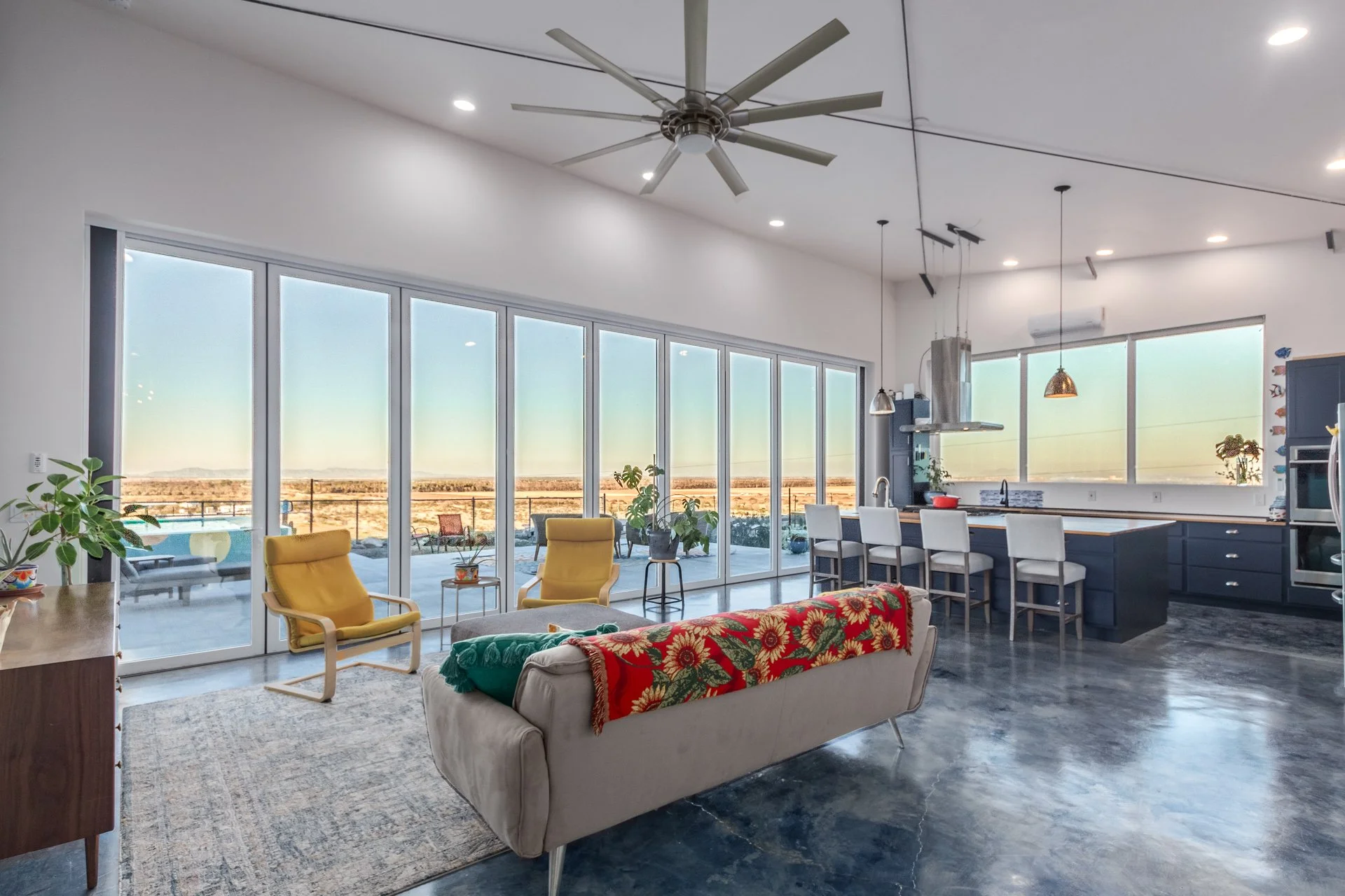 Open-concept kitchen and living room with large windows showing a desert landscape, ceiling fan, yellow armchairs, sofa with floral blanket, kitchen island with white chairs, and modern appliances.
