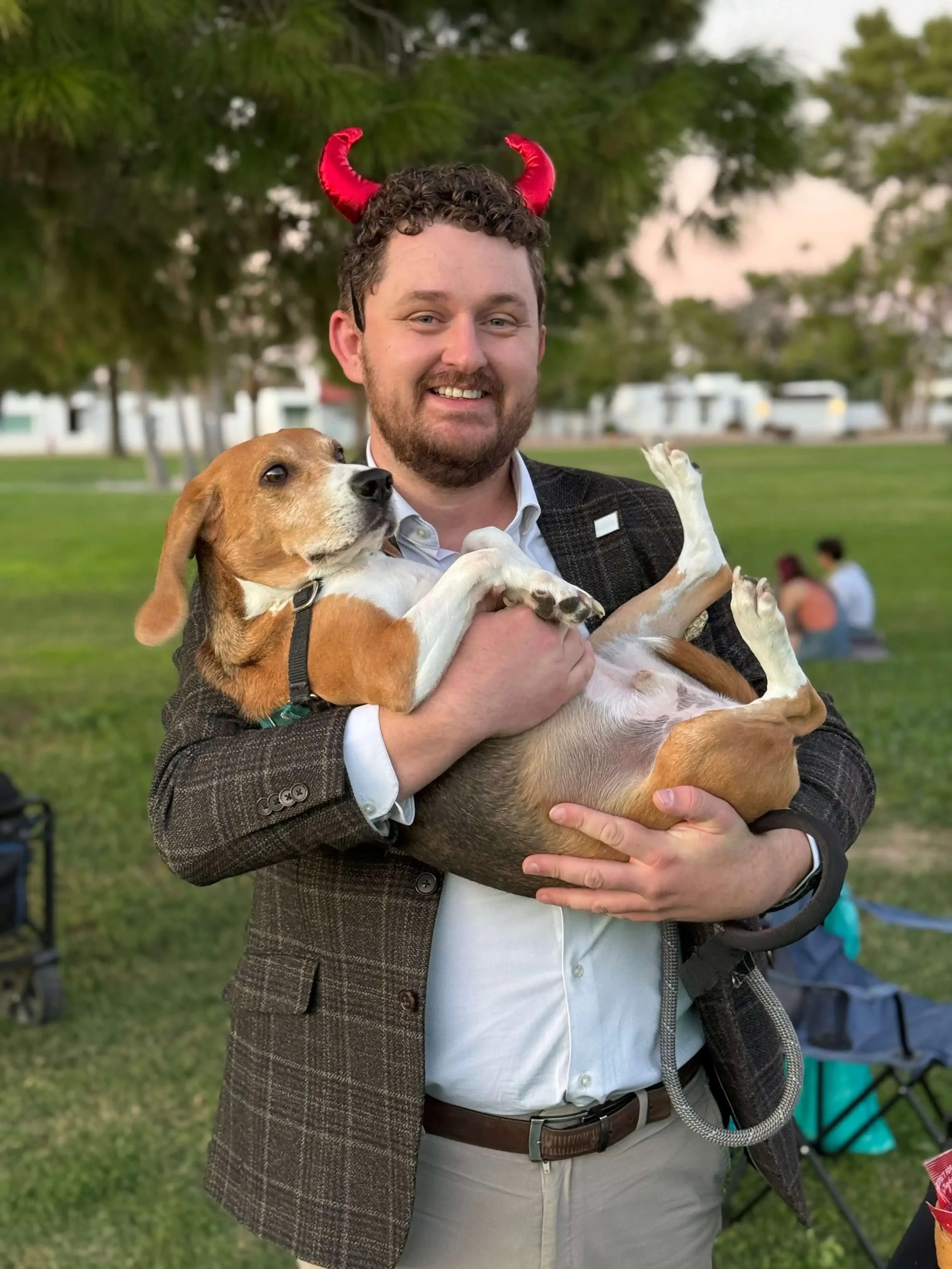 A smiling man in a blazer and red devil horns holding a beagle dog like a baby at the Bryant Real Estate Mutts and Munchkins outdoor Halloween event in Phoenix.