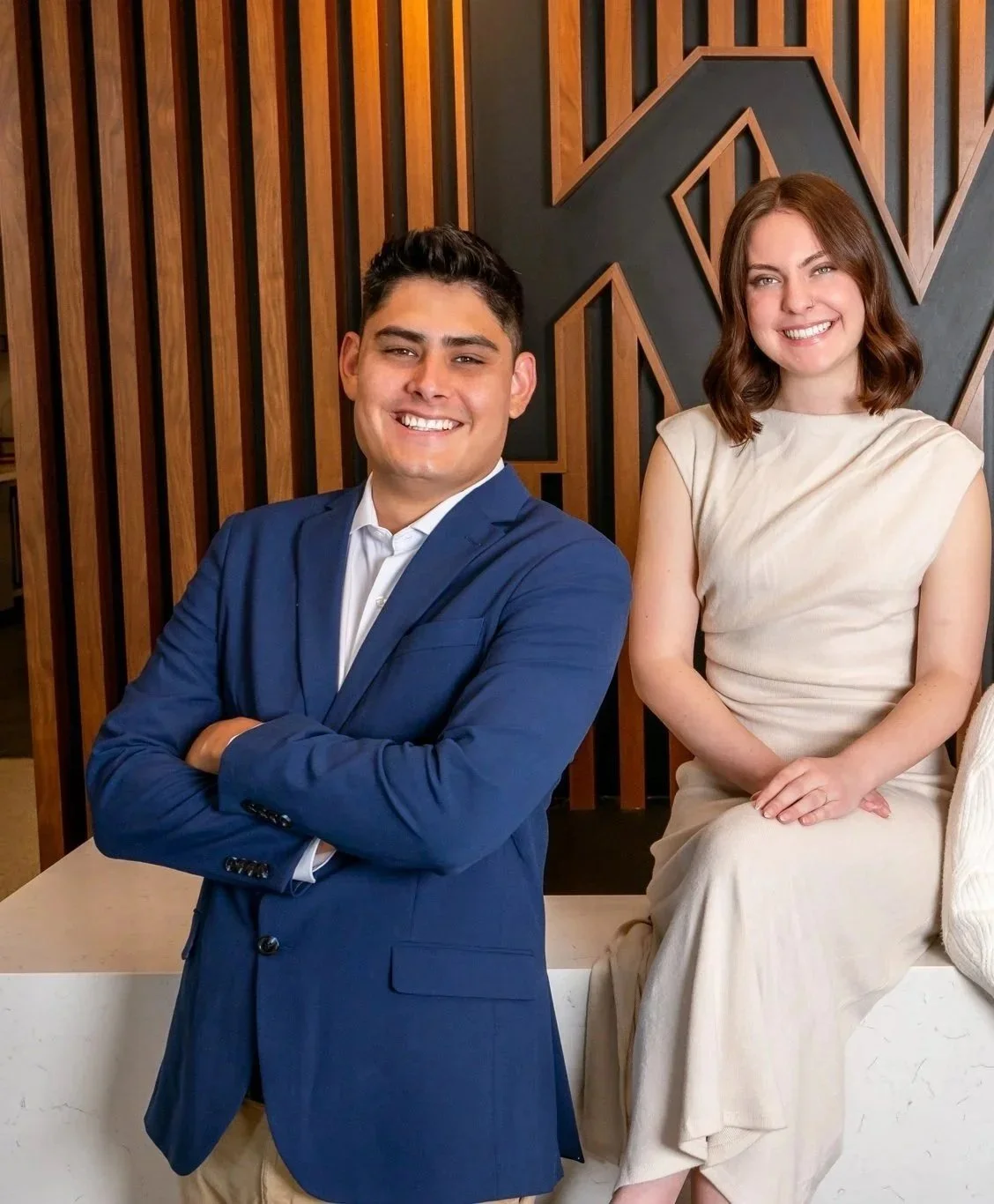 Smiling man in a blue blazer with arms crossed standing next to a seated woman in a cream dress, both in a modern indoor setting with wood-paneled wall behind them.