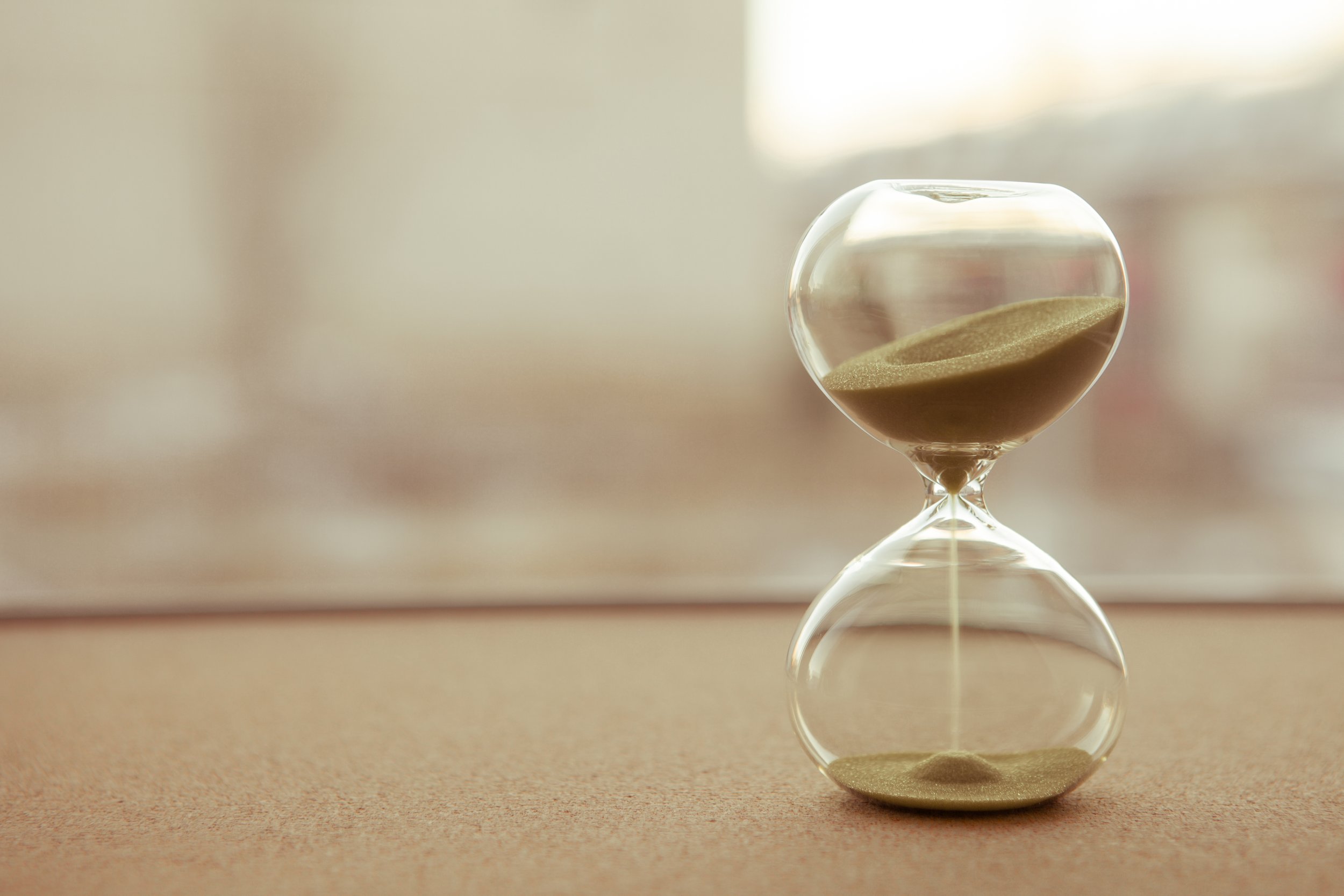 An hourglass filled with sand, with the top half almost full and the bottom half nearly empty, sitting on a flat surface with a blurred background.