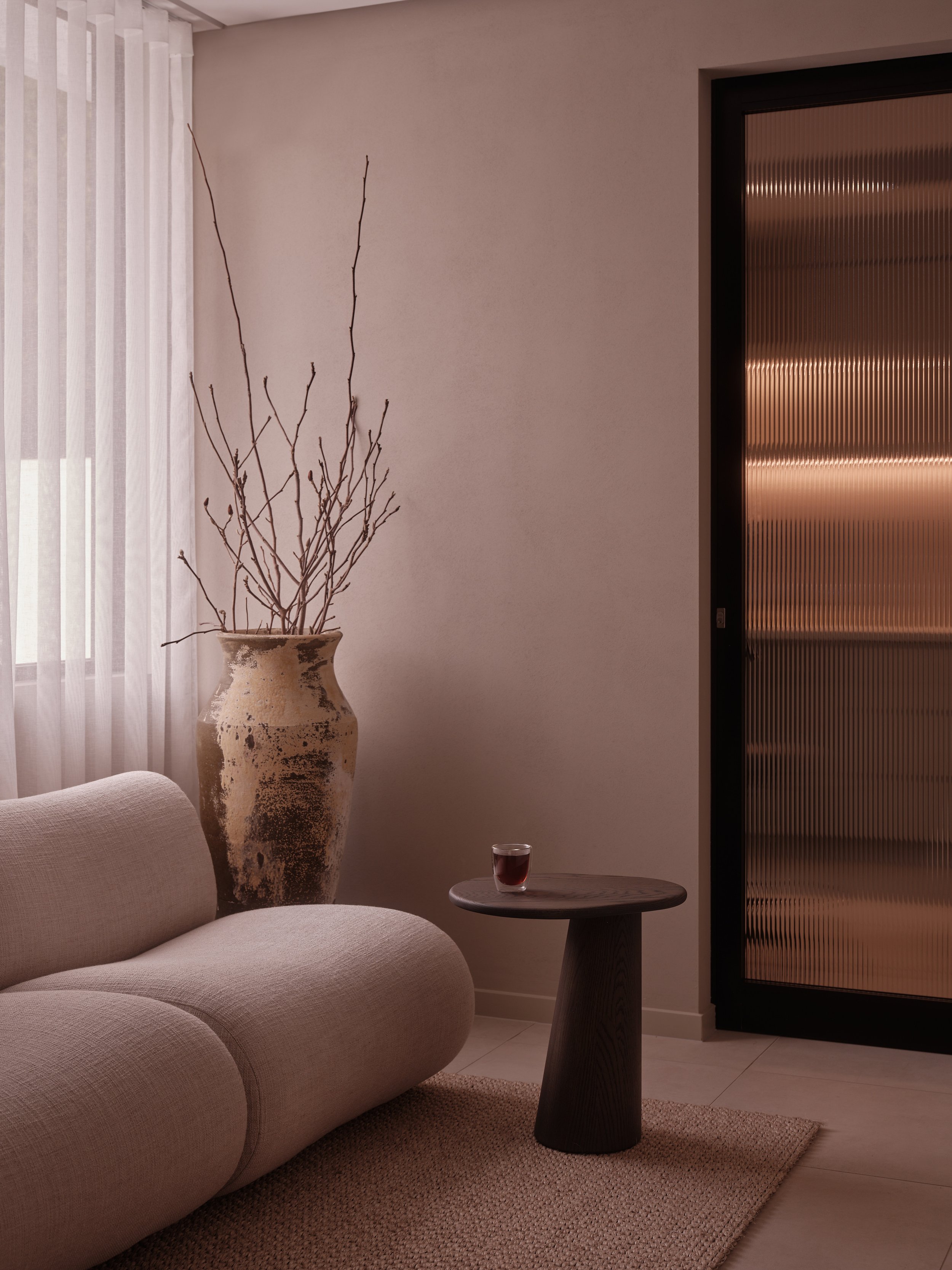 A minimalist living room corner with a beige sofa, a large decorative vase with bare branches, a small round black side table with a glass of red wine, a window with sheer curtains, and a textured sliding door with warm lighting behind.