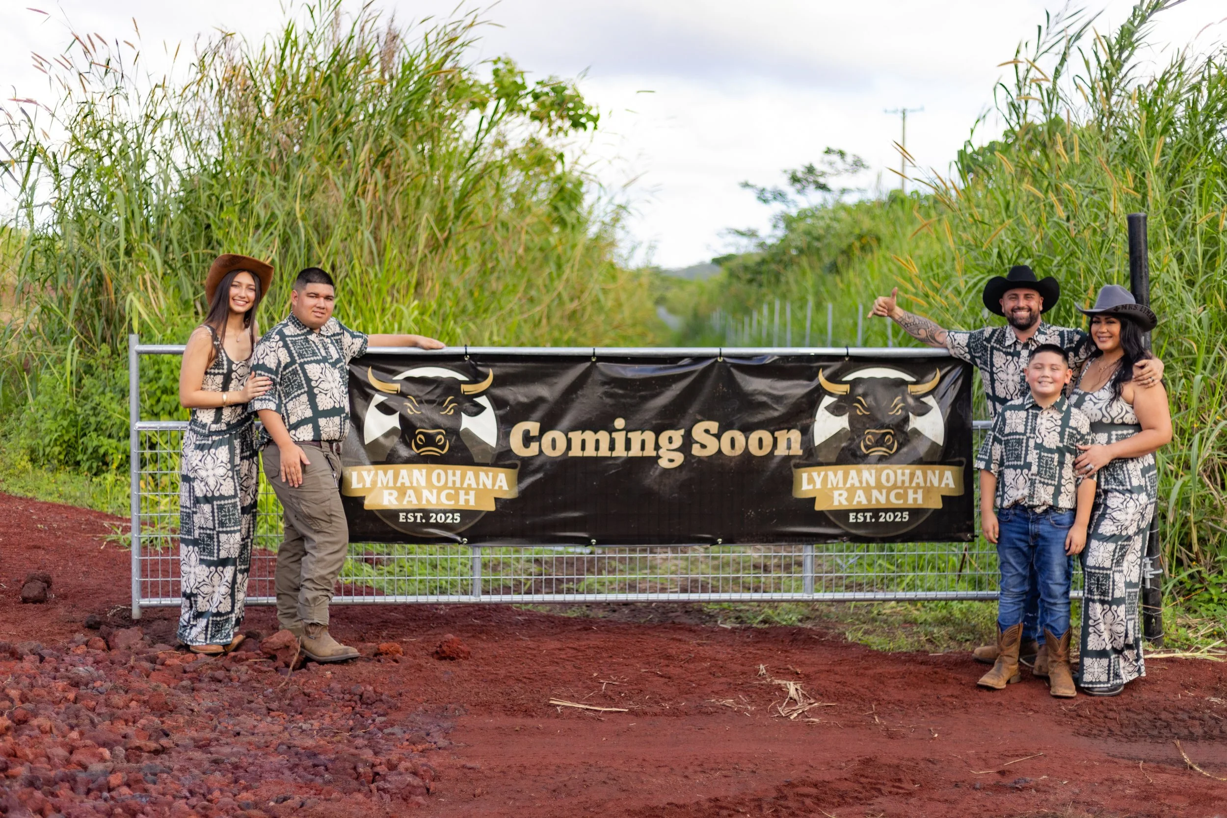 Family of four standing on red dirt road in front of a black banner with a bull logo, text 'Coming Soon Lyman Ohana Ranch EST. 2025,' flanked by tall green grass and trees, with partly cloudy sky overhead.