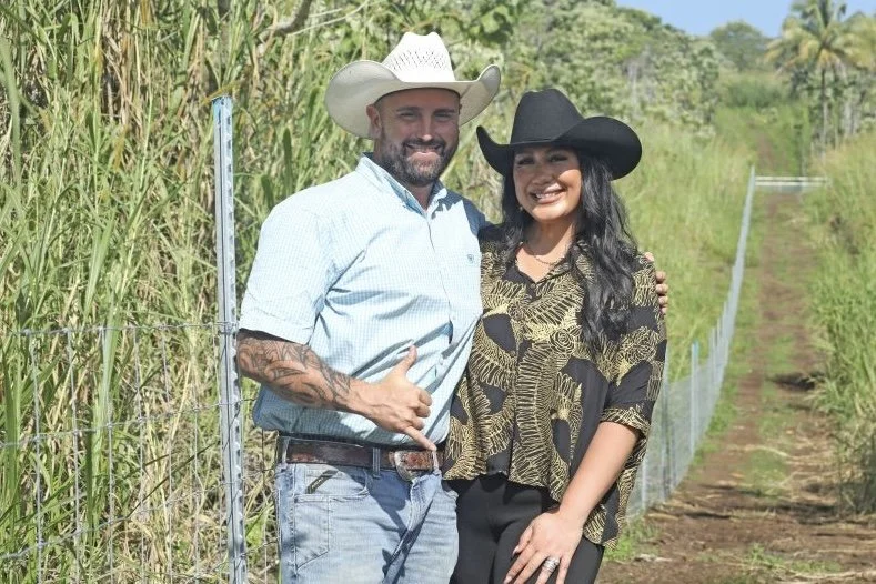 A man and woman in cowboy hats standing outdoors by a wire fence, smiling at the camera, surrounded by green plants and trees.