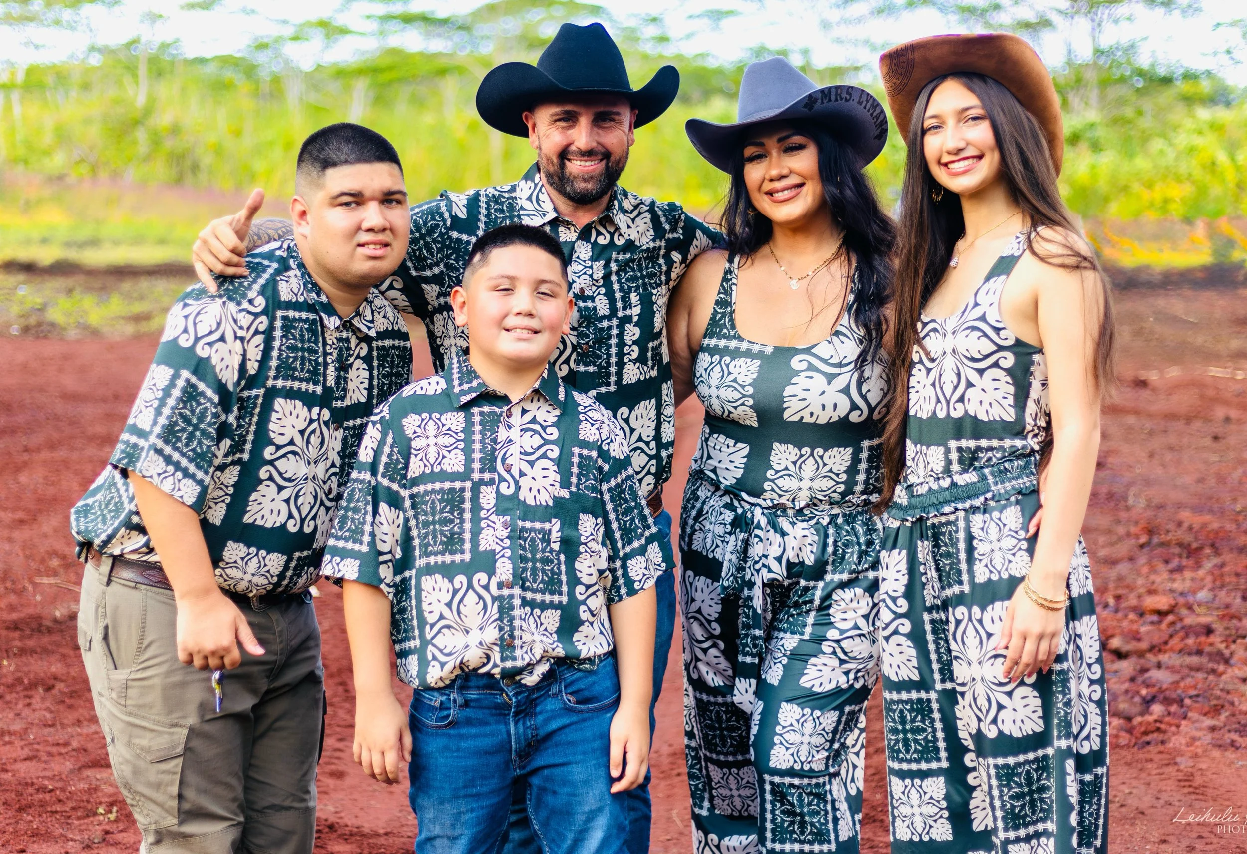 A family of six members dressed in matching blue and white patterned outfits posing outdoors on red soil with green foliage in the background. The adults are wearing cowboy hats, and the family members are smiling at the camera.