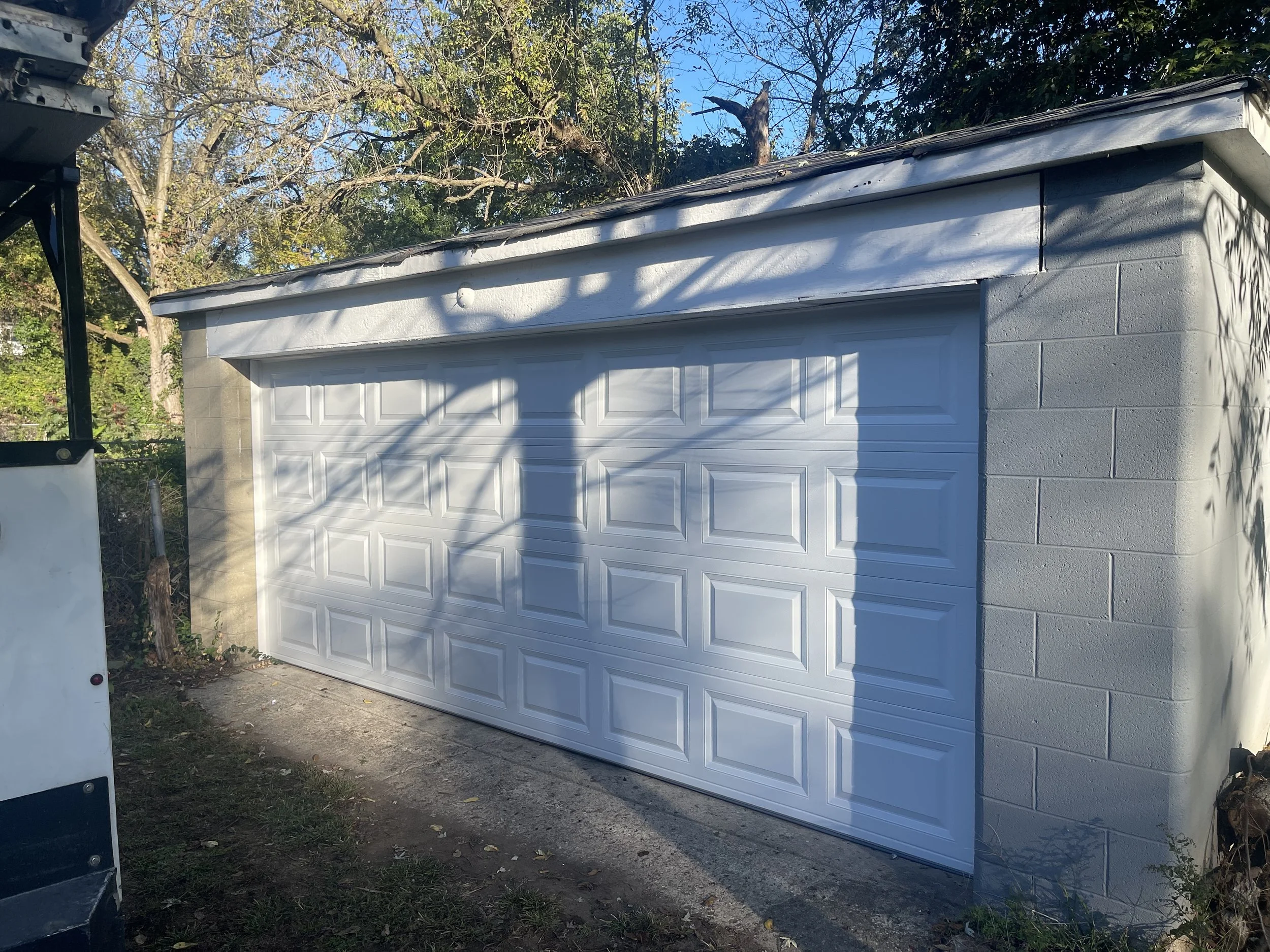 White 18 x 8 foot garage door set into the side of a tan building surrounded by trees