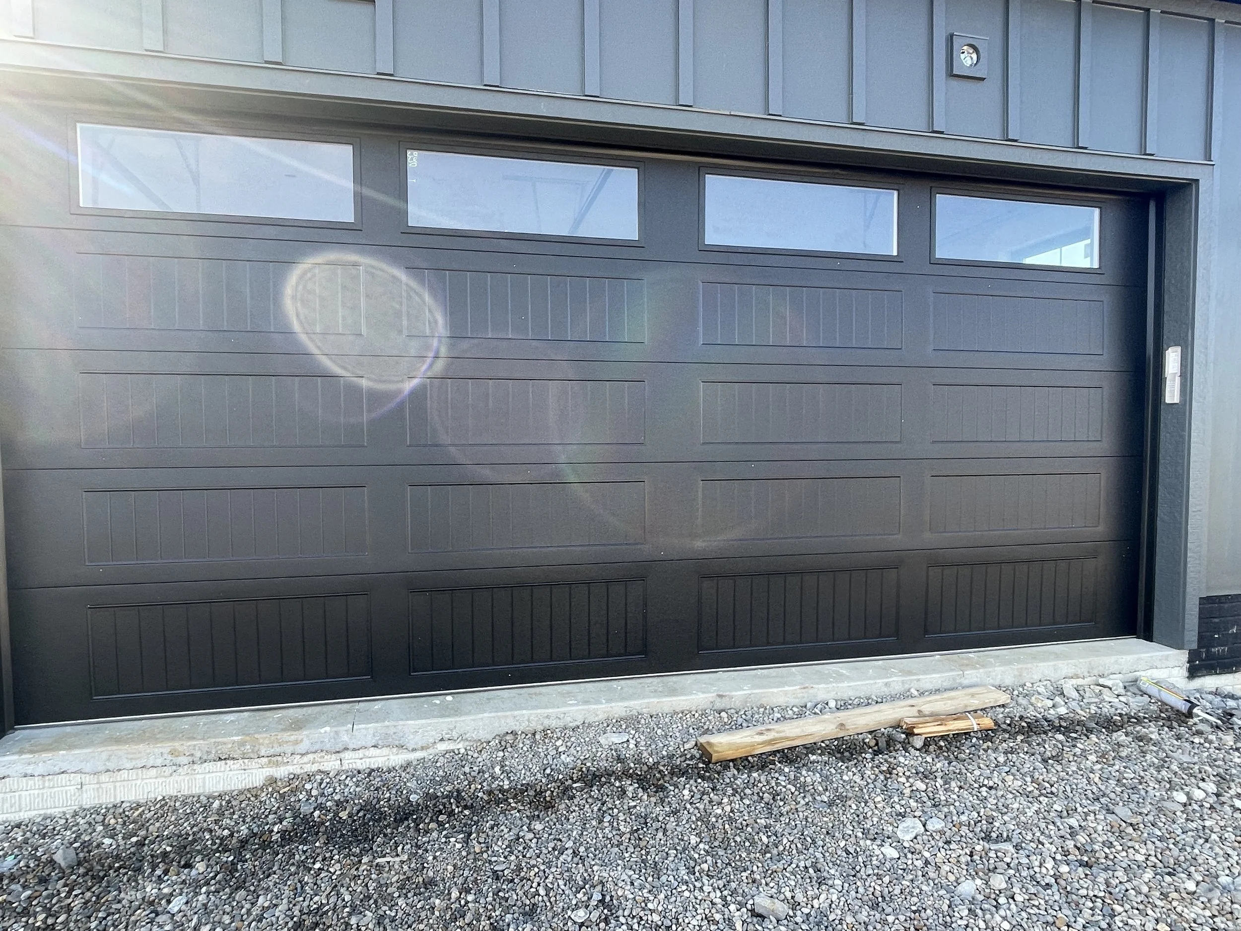 A gray 18 by 7 garage door with windows in the top section set into the side of a gray steel wall.