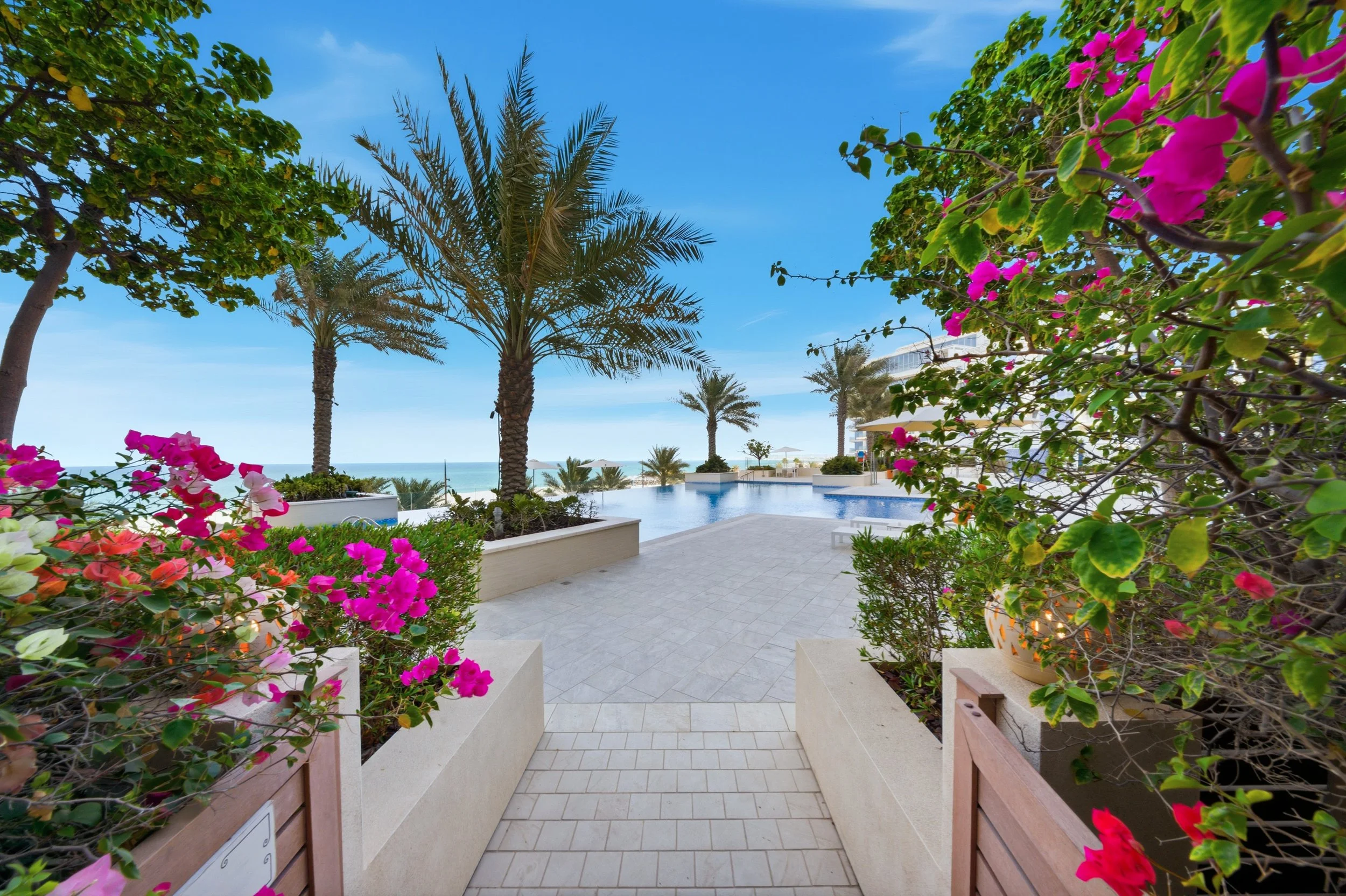 Tropical resort pool area with palm trees, flowering plants, and ocean view under a partly cloudy sky.