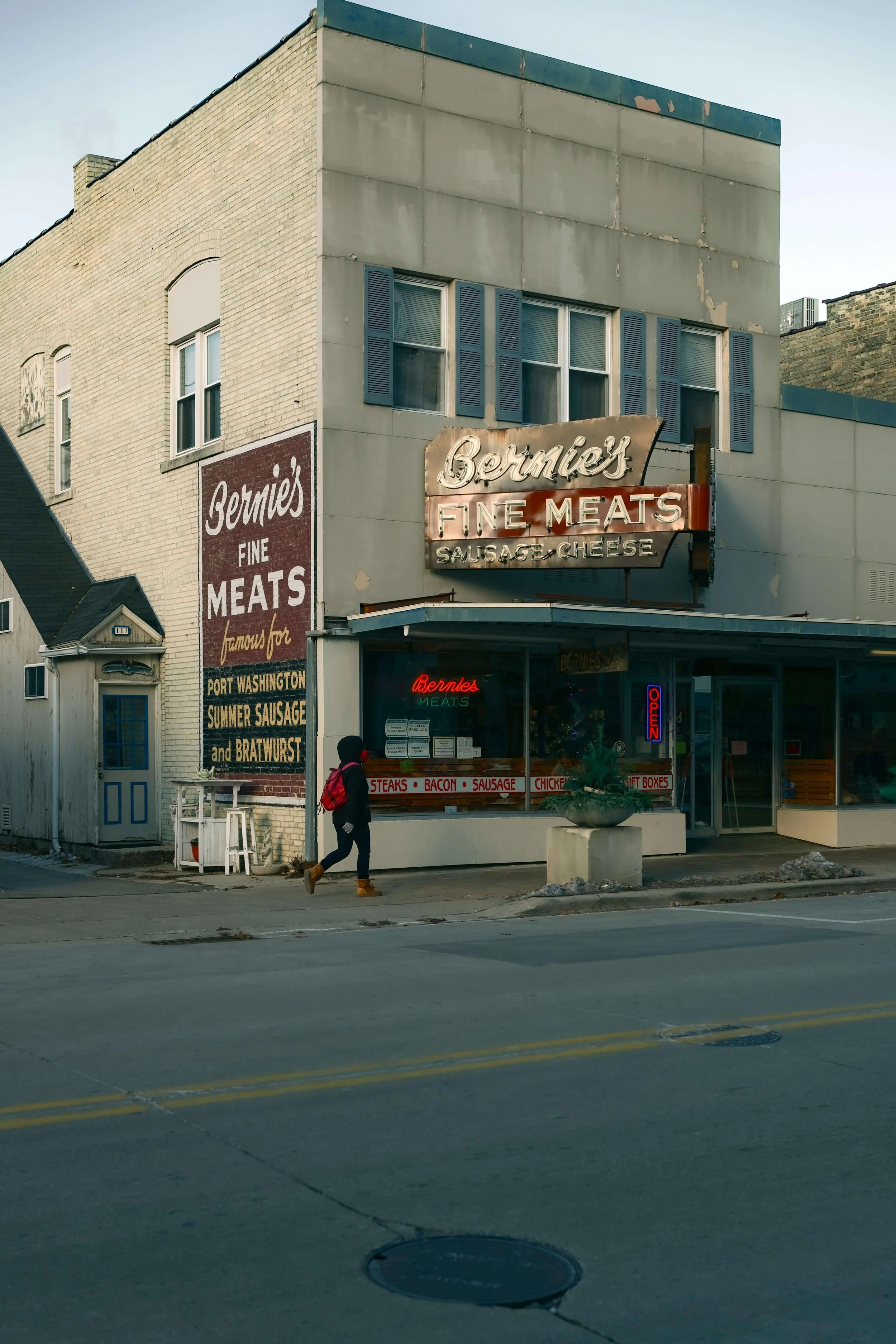 Old-fashioned storefront of Bernie's Fine Meats with neon signage, a sidewalk, and a pedestrian walking past, featuring signs advertising meats like steaks, bacon, sausage, chicken, and gift boxes, in an urban area.