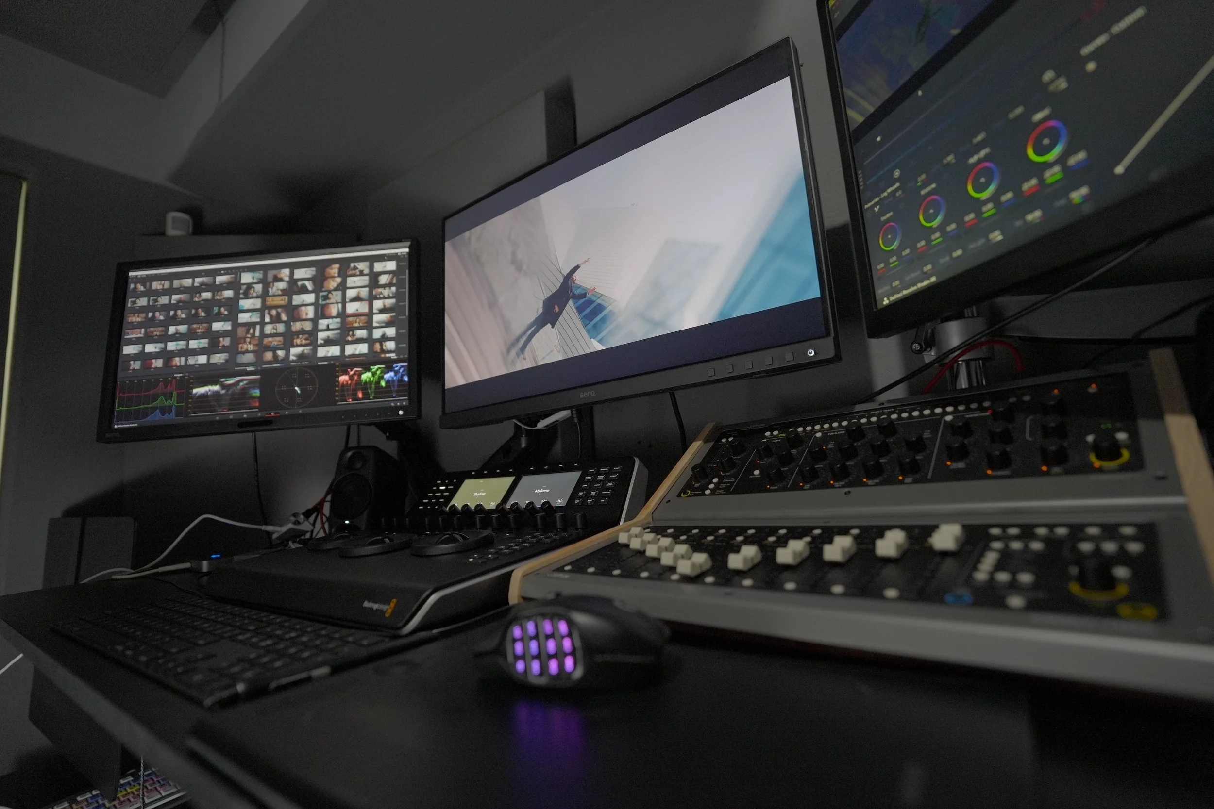 A professional video editing setup with three monitors, editing software, a keyboard, a mouse, and audio equipment on a desk.