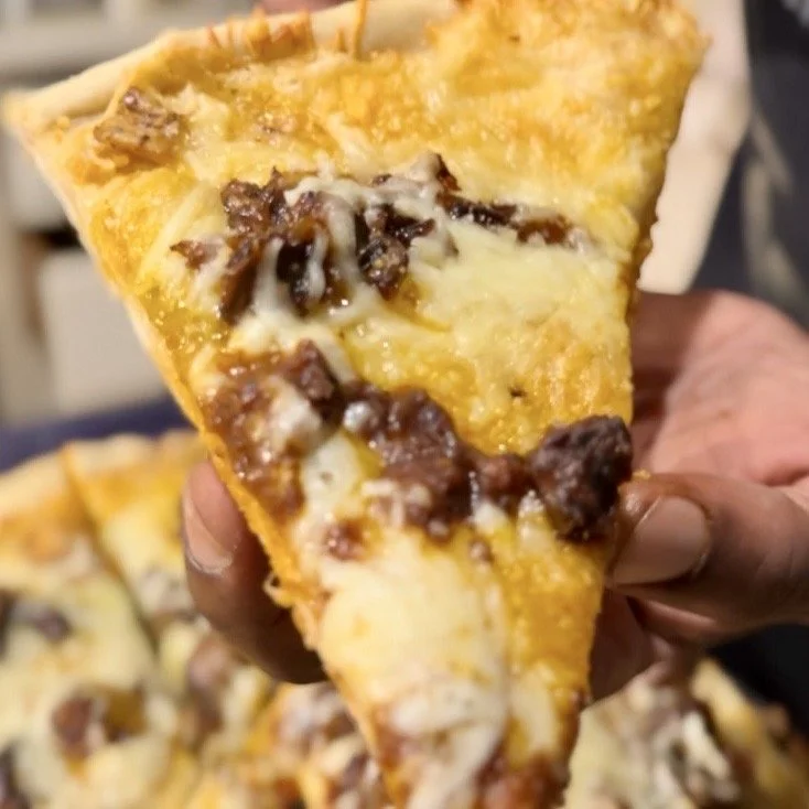Close-up of a hand holding a slice of oxtail pizza with melted cheese, oxtail, and Ed's special sauce.