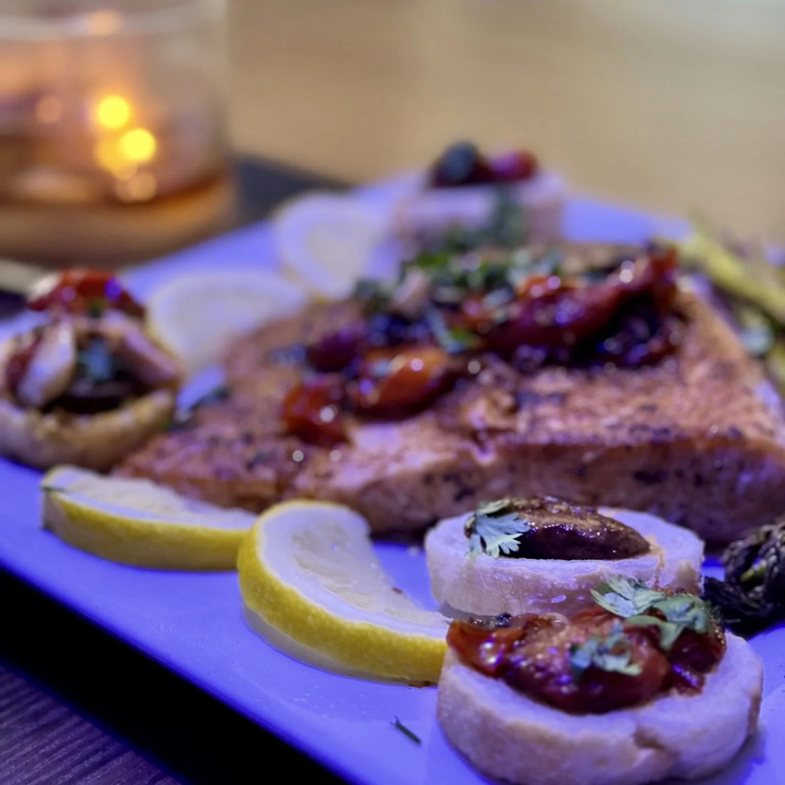 Close-up of a platter with lemon slices, toasted bread topped with a spread and garnished with herbs, grilled vegetables, and a piece of grilled fish with sauce and herbs. In the background, a candle in a glass holder.
