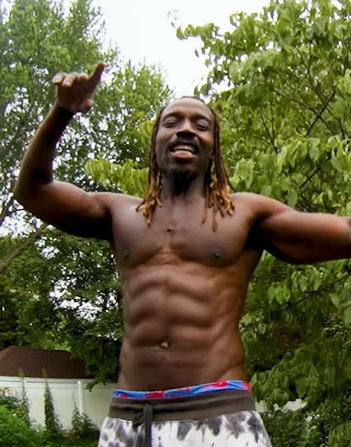 A shirtless man with dreadlocks smiling outdoors among green trees.