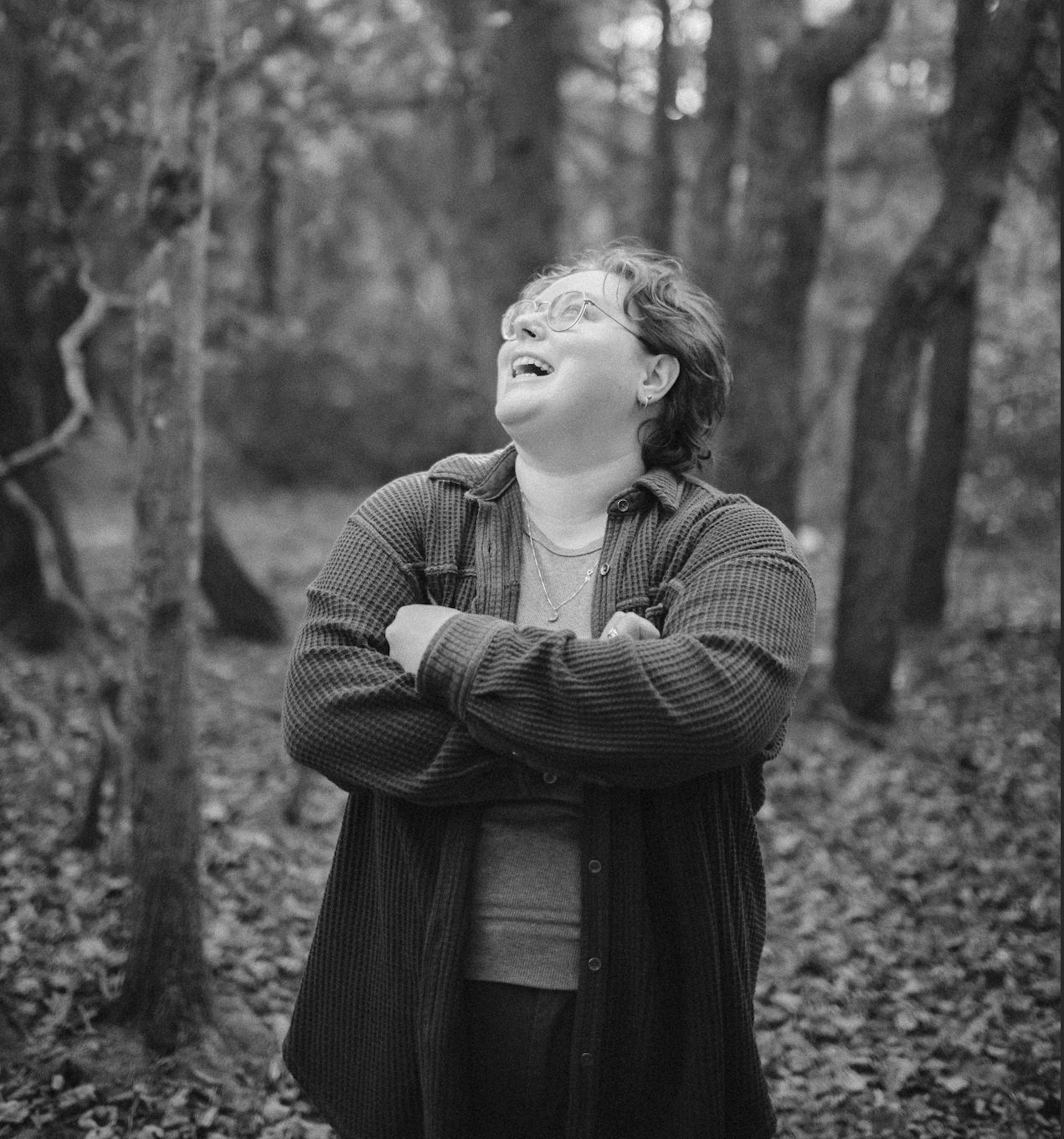 A woman smiling with her arms crossed in a forest setting, wearing glasses, earrings, layered necklaces, and a textured jacket.