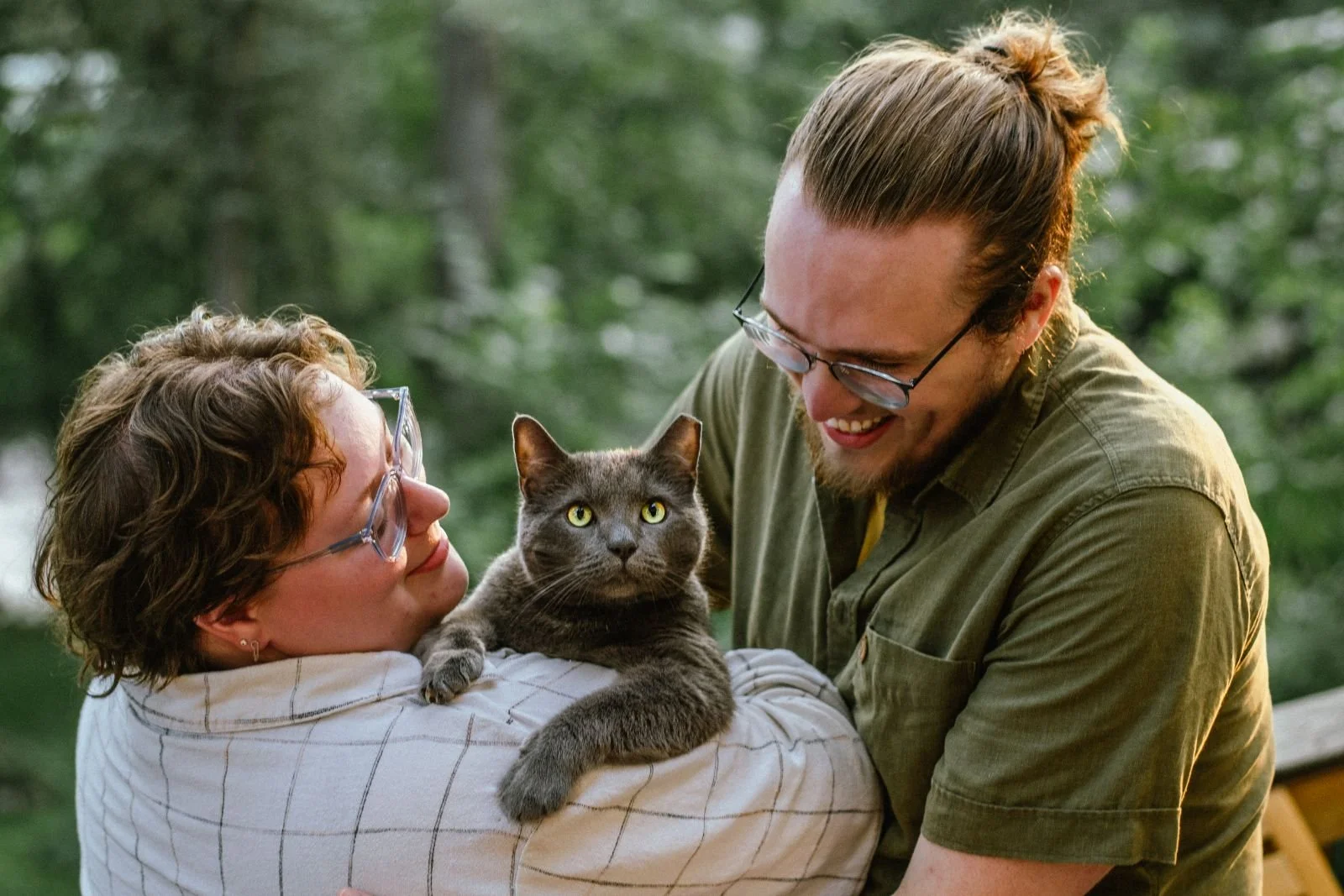 A man and woman wearing glasses are holding a gray cat with yellow eyes outdoors in a green wooded area, smiling at each other.