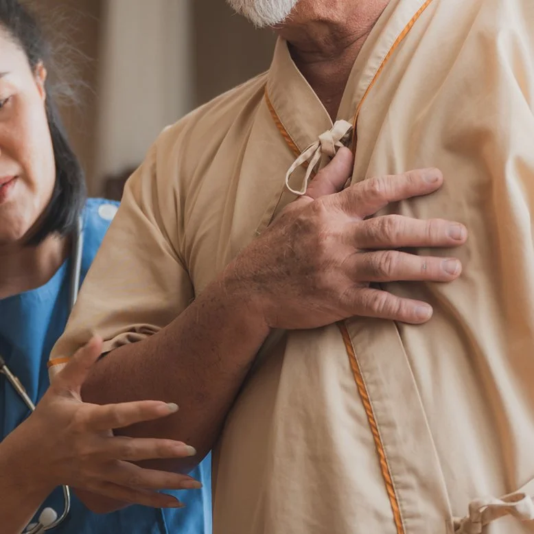 A person wearing a hospital gown placing their hand on their chest, being assisted by a healthcare worker in blue scrubs.