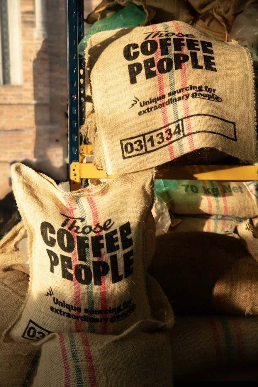 Bags of coffee beans labeled "Choose COFFEE PEOPLE," hanging on a shelving unit in a warehouse. The bags are made of burlap and have black and red text, with numbers printed on them.