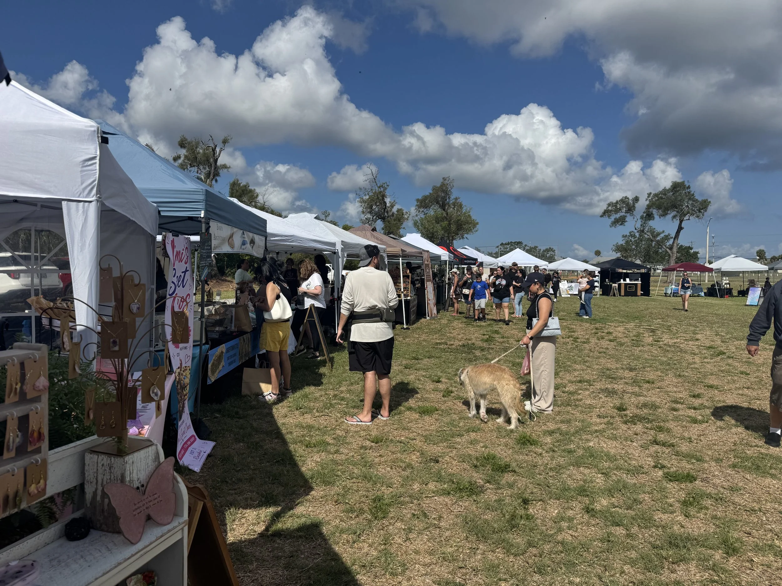 An outdoor market or fair with multiple vendor tents under a partly cloudy sky. People, including a woman walking a dog, browse the stalls on a grassy field with trees in the background.