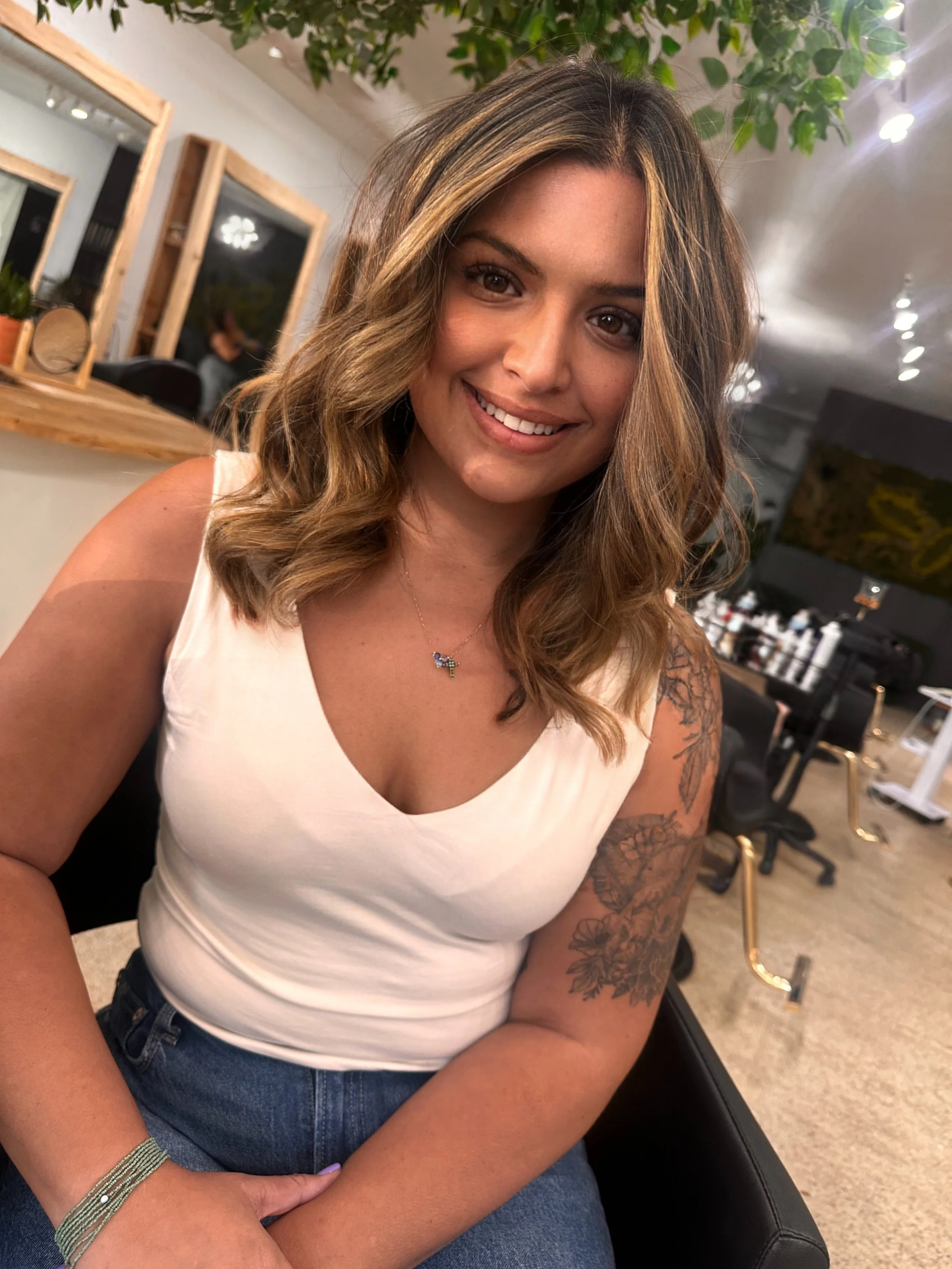 A woman with shoulder-length wavy hair, wearing a white sleeveless top and blue jeans, smiling at the camera in a salon or hair studio with mirrors and salon equipment in the background.