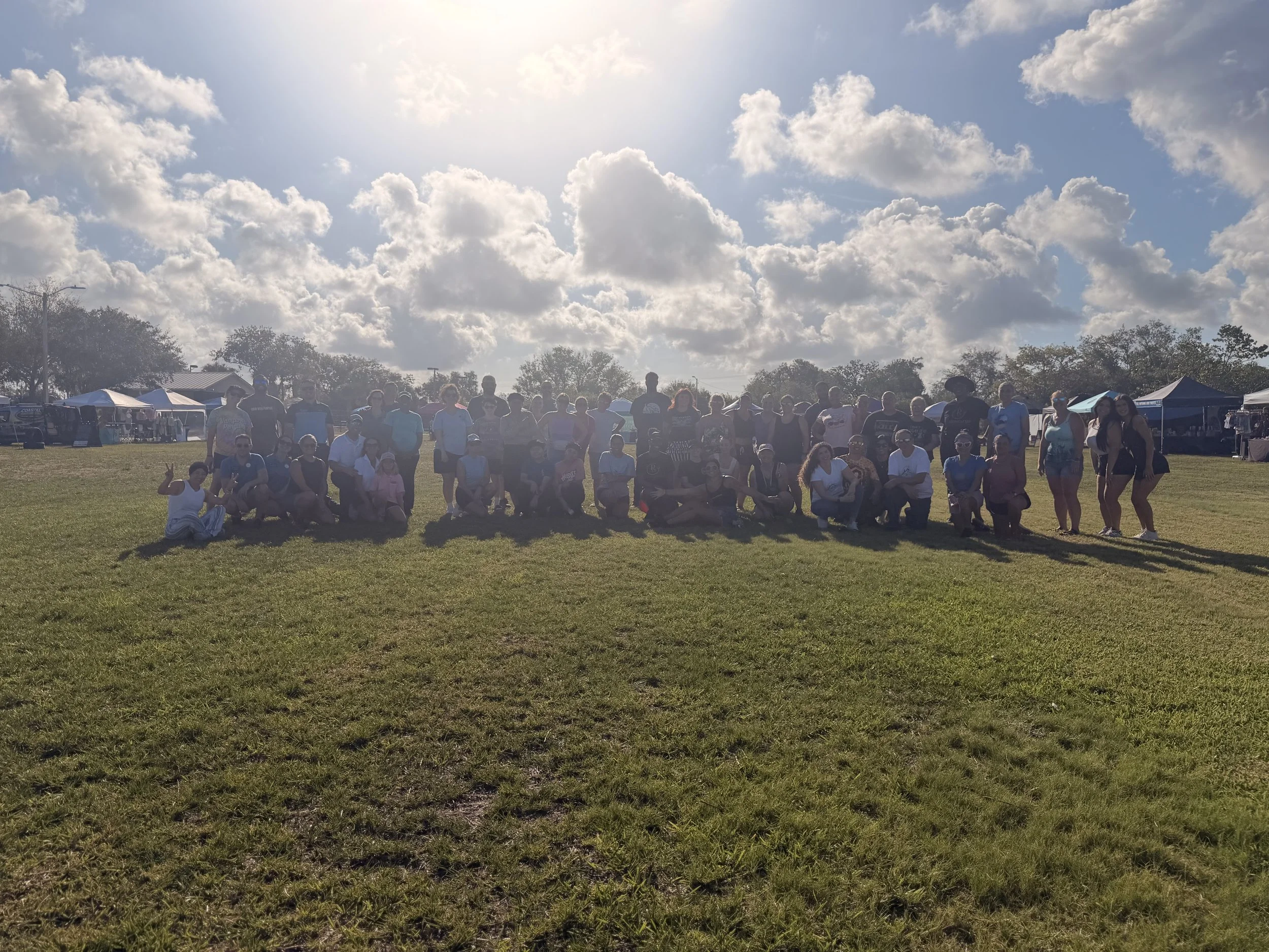 Group of people standing and sitting on grass field during the daytime with tents and trees in the background under partly cloudy sky.