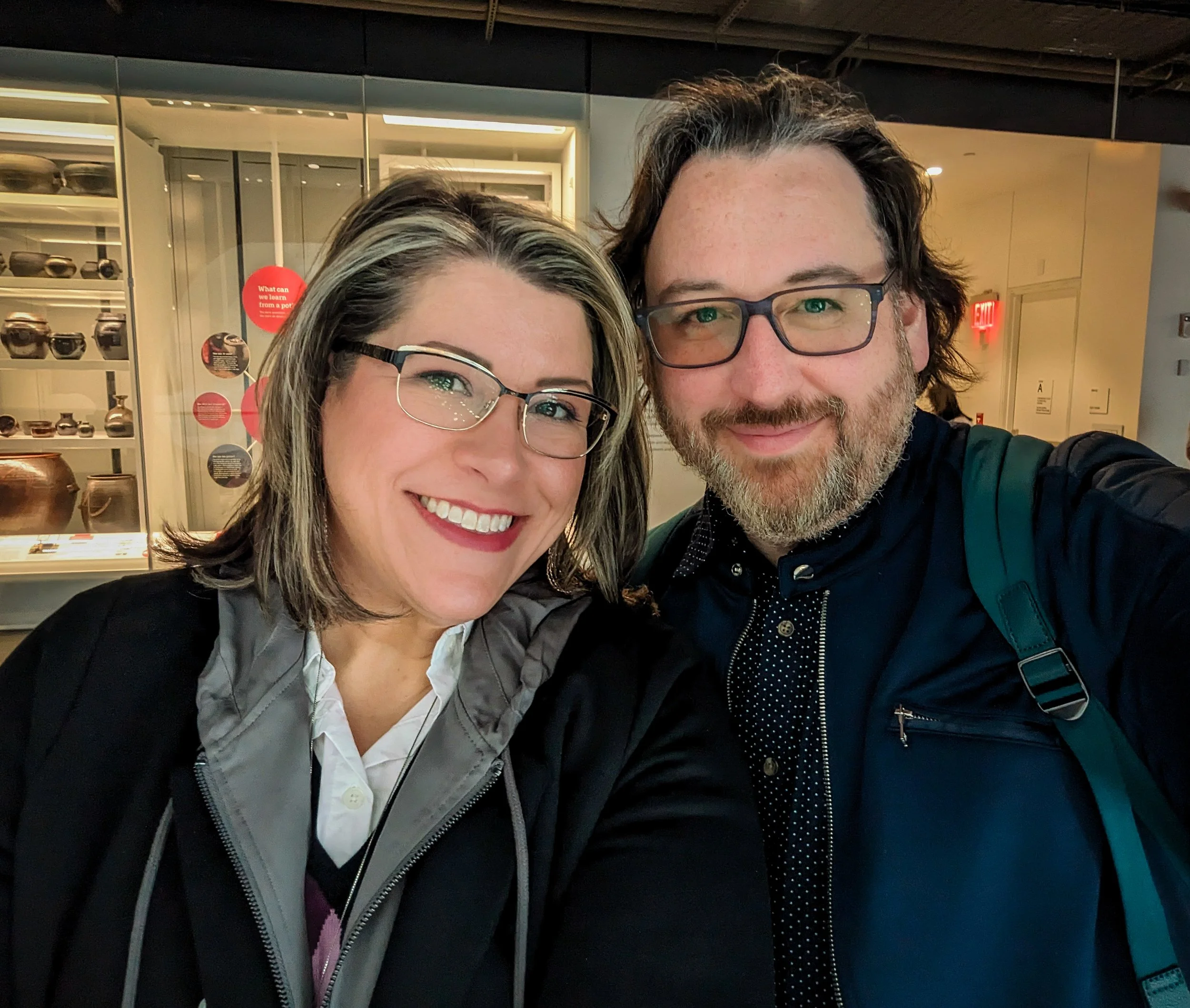 A smiling woman and man taking a selfie together in an indoor setting. The woman has shoulder-length hair, glasses, and is wearing a white shirt with a jacket. The man has dark hair, glasses, a beard, and is wearing a dark shirt with a backpack strap visible. In the background, there is a display case with pottery and informational signs.