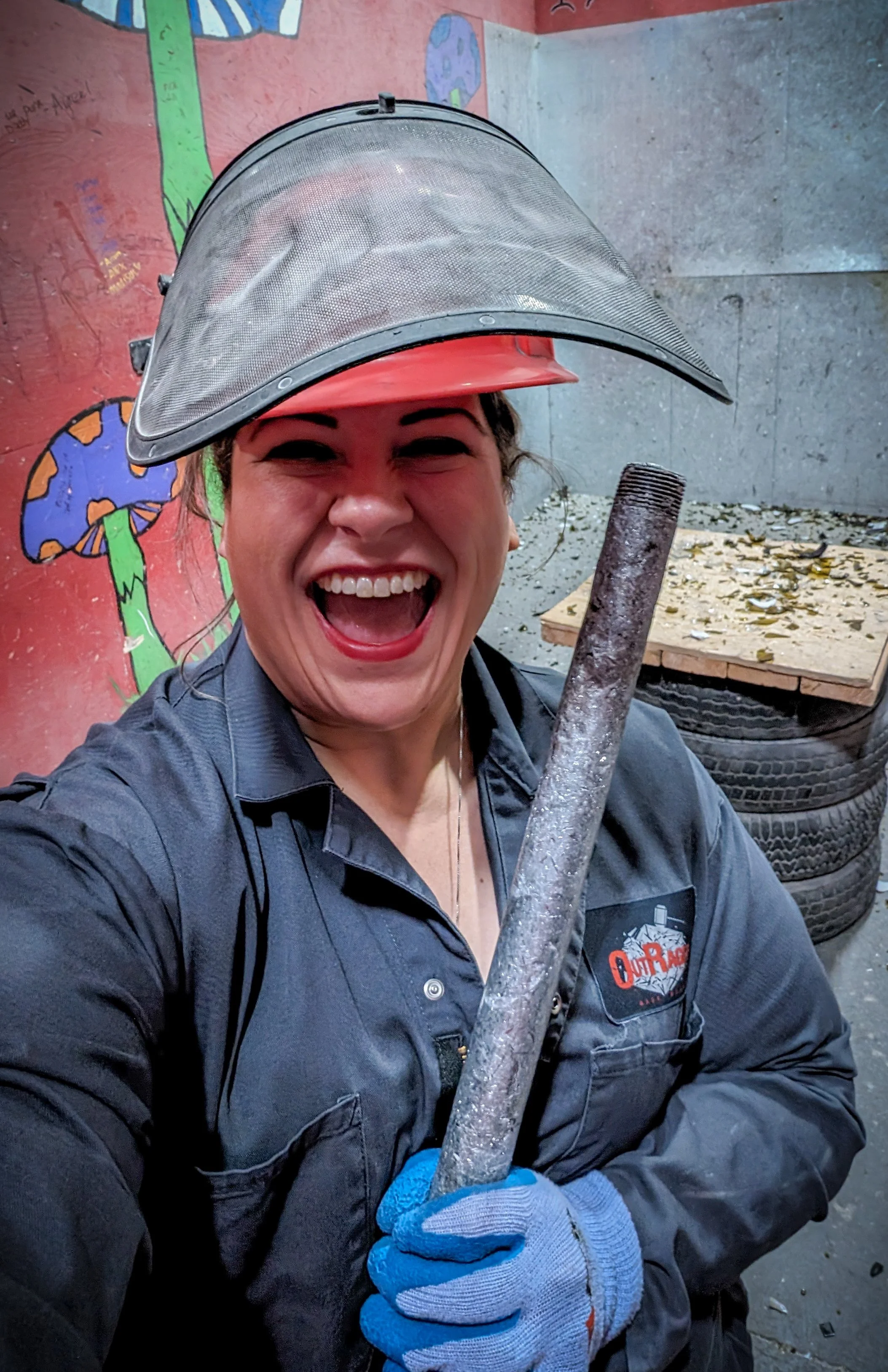 A woman in a black work uniform, wearing gloves and protective helmet, smiling excitedly while holding a metal pipe at a workshop or garage with tires and painted mural in the background.
