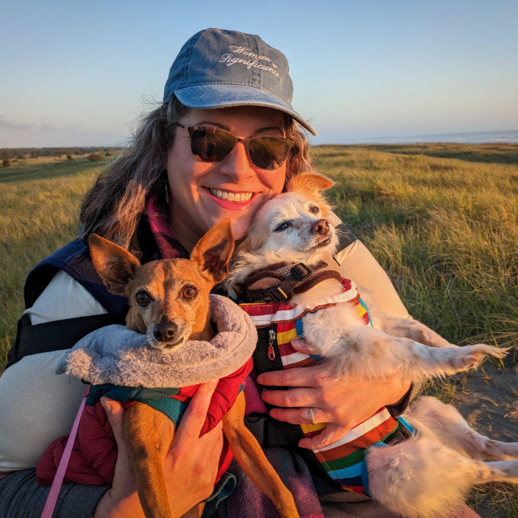 A smiling woman wearing sunglasses and a blue cap holding two small dogs in a grassy field during sunset.