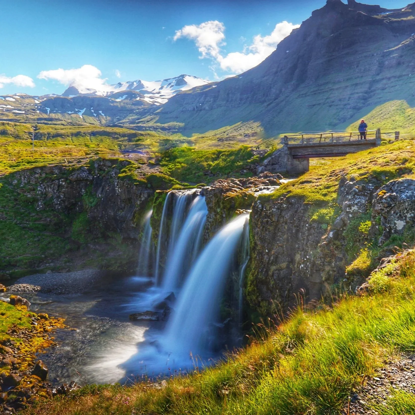 A scenic landscape featuring a waterfall with flowing water, green foliage, a mountain range with snow-capped peaks, and a blue sky with clouds. A person stands on a small bridge above the waterfall.