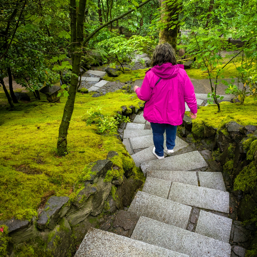 Person in a pink jacket walking on a stone pathway through a green, moss-covered garden.