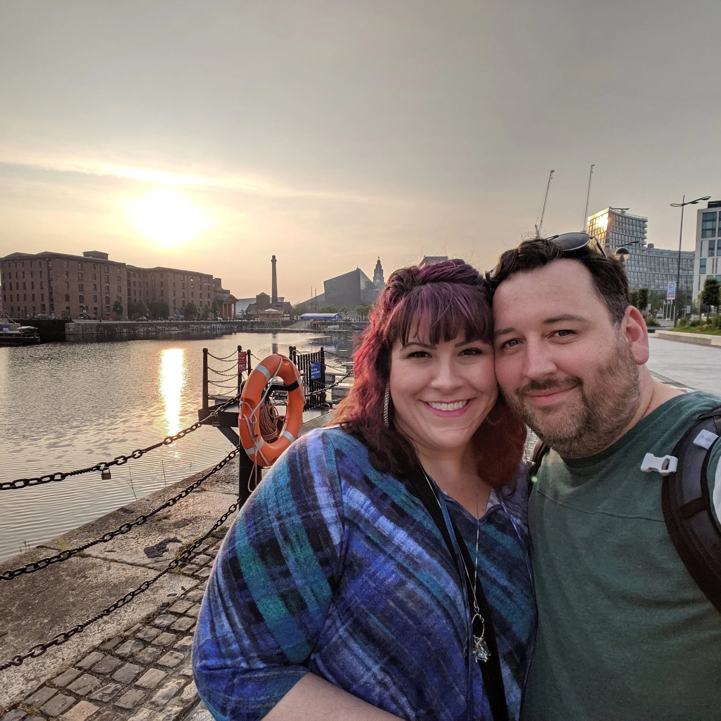 A smiling couple taking a selfie by a waterfront at sunset. The woman has purple hair and is wearing a blue plaid shirt; the man has dark hair and a beard, wearing a green shirt with a backpack. City buildings and a dock with a life buoy are visible in the background.