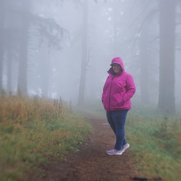 Woman wearing a pink waterproof jacket and jeans walking on a foggy forest trail.