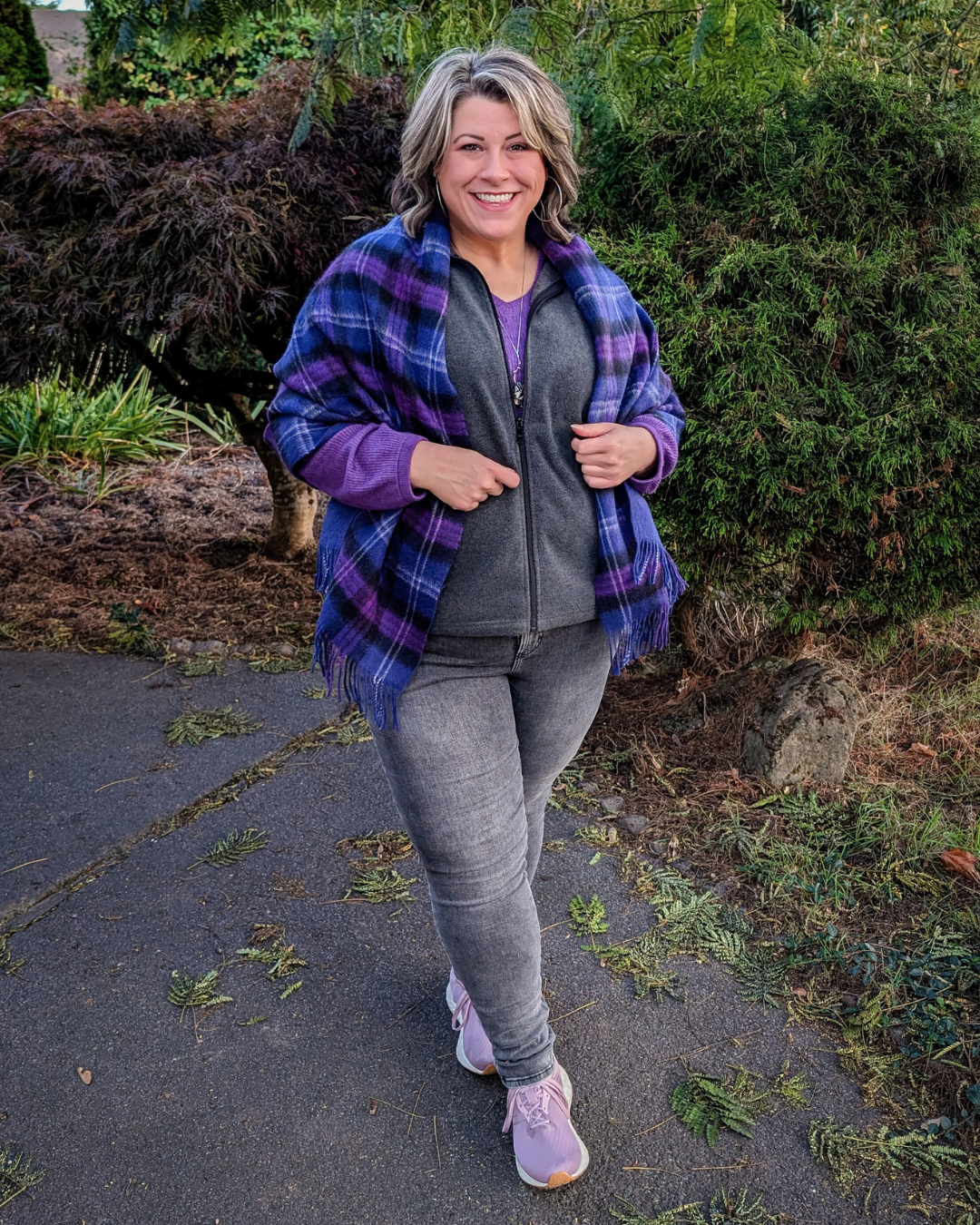 A woman with short blonde hair smiling and wearing a purple and blue plaid shawl, gray zip-up hoodie, gray jeans, and pink sneakers, standing on a sidewalk with greenery and bushes in the background.