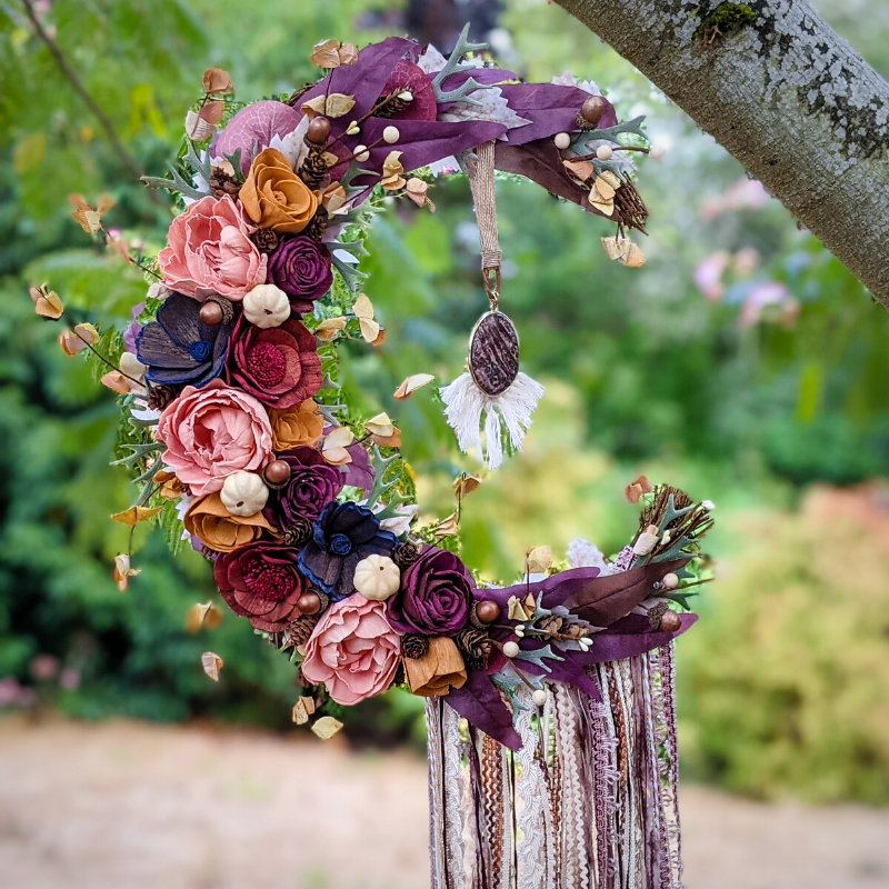 Playful moon-shaped floral wreath decorated with artificial flowers, leaves, and a hanging tassel, hanging from a tree branch in an outdoor setting.