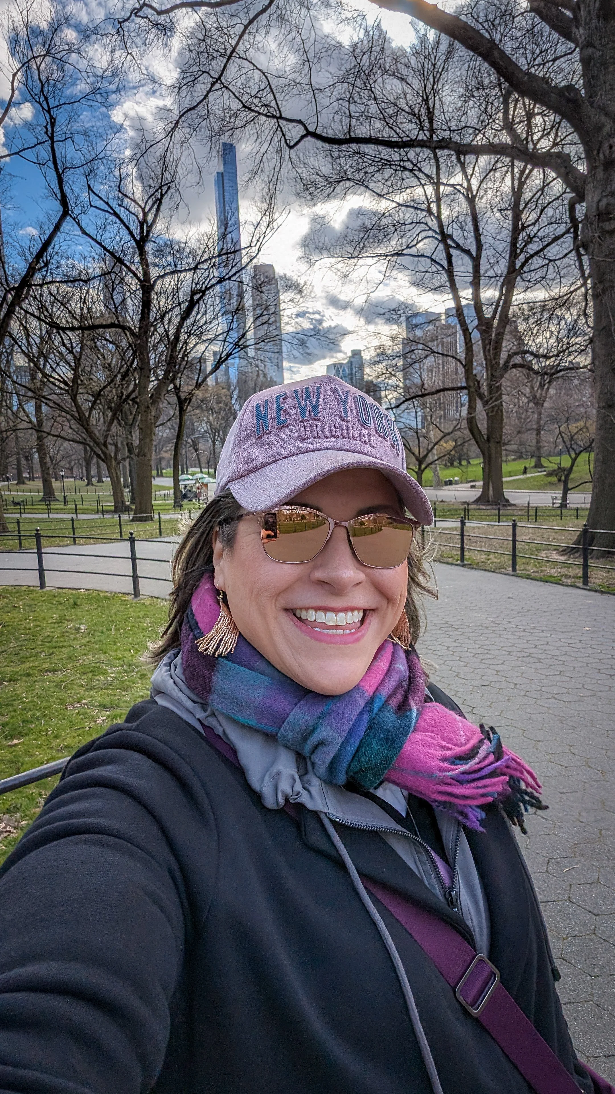 A woman smiling in a park with trees and city skyscrapers in the background, wearing sunglasses, a pink cap that says 'New York Original,' a colorful scarf, and a dark jacket.