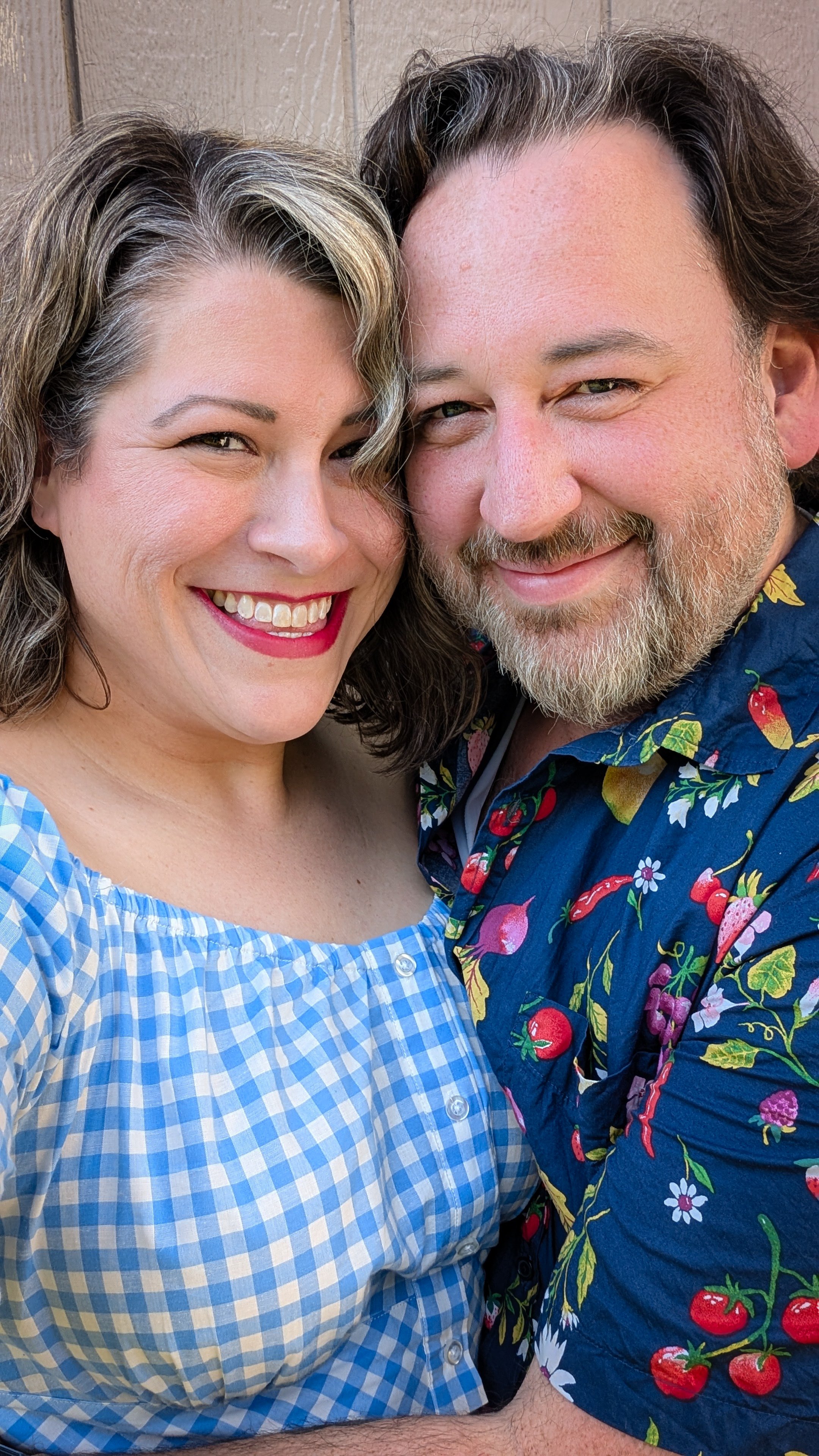 A smiling couple taking a close-up photo outdoors, with the woman in a blue checkered dress and the man wearing a colorful Hawaiian shirt, standing closely together.